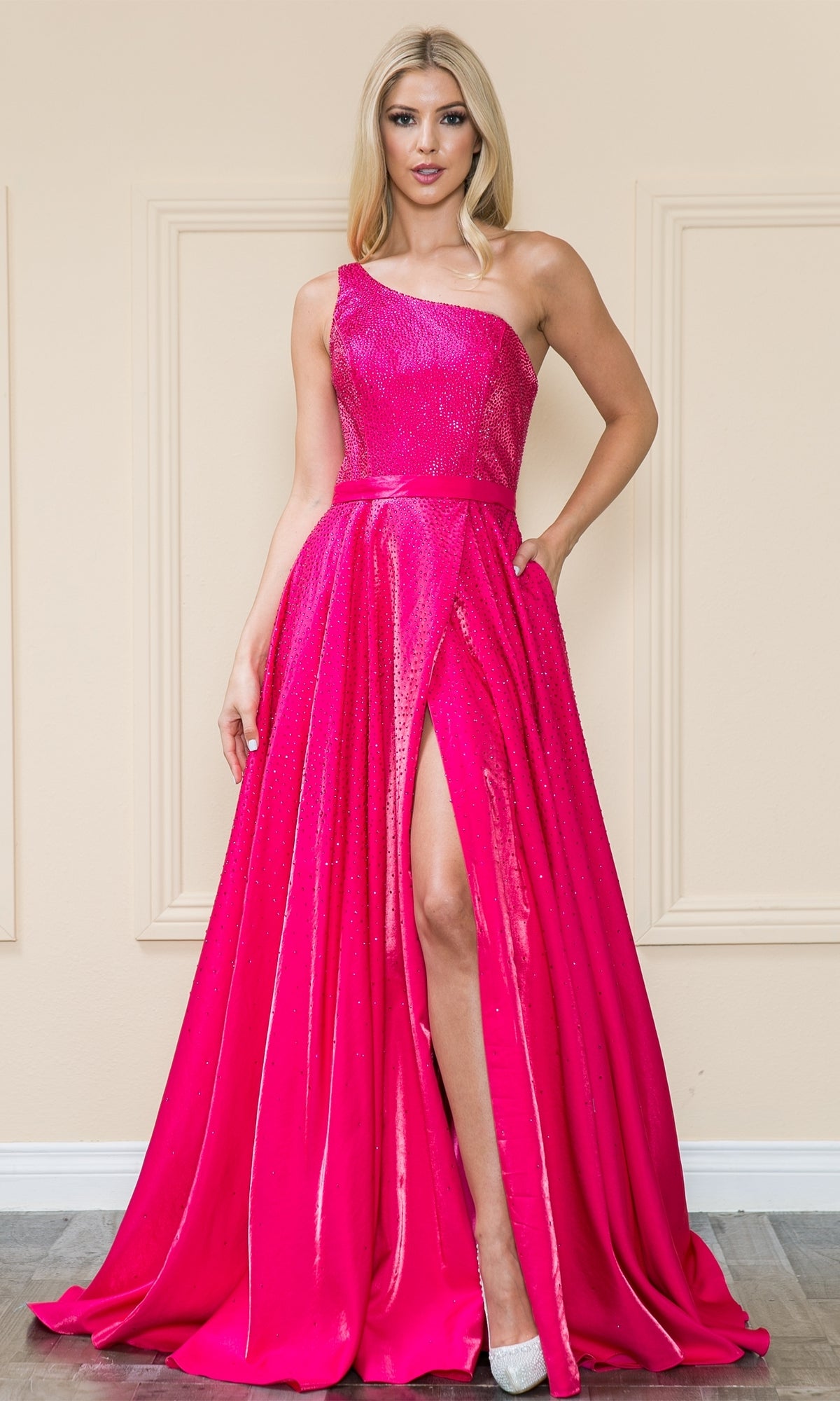 A woman stands against a beige wall wearing the Poly USA 8914, a hot pink one-shoulder long A-line prom gown.