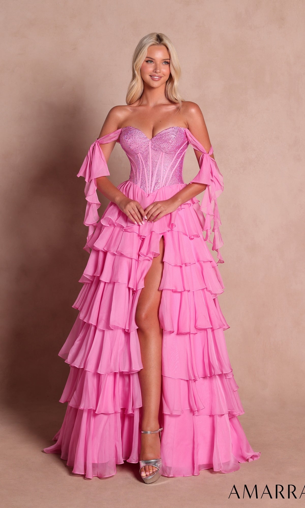 A woman wearing the Long Prom Dress: Amarra 89285 in bright pink, featuring a ruffled off-the-shoulder design, an embellished corset bodice, and a high slit, stands against a beige background.