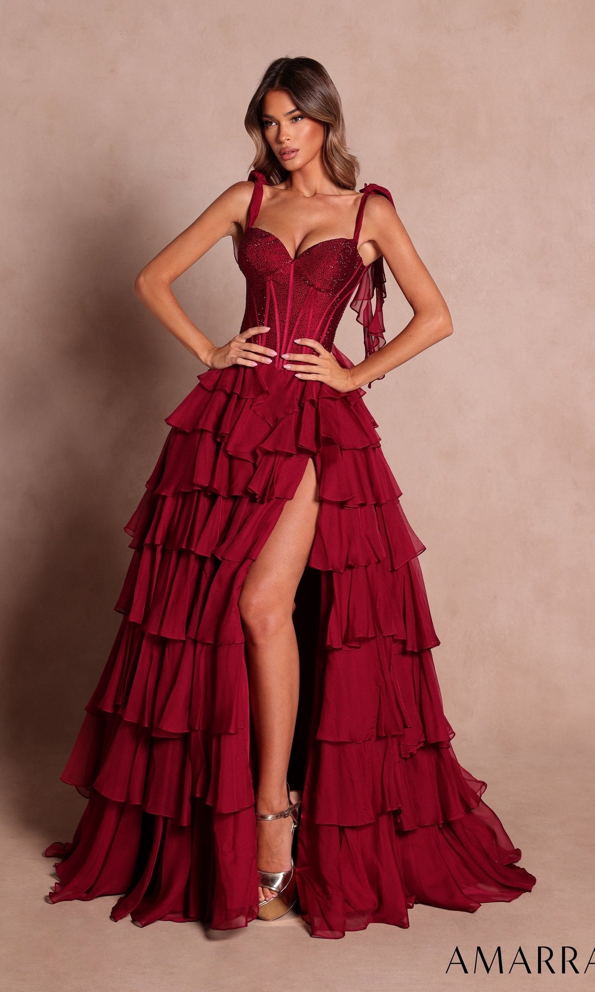 A woman in the Amarra 89285 long prom dress in wine, featuring a sleeveless ruffled gown with a high slit and an embellished corset bodice, stands against a neutral background.