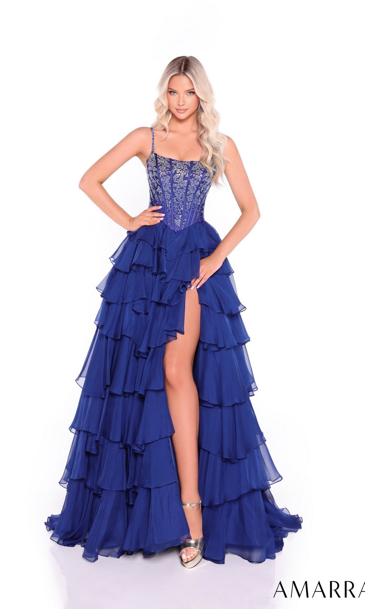 A woman poses in the Lace-Up Long Tiered Prom Dress: Amarra 89459, featuring royal blue tiers, an embellished corset-style bodice, and a high leg slit. She pairs it with silver heels and stands against a white background.