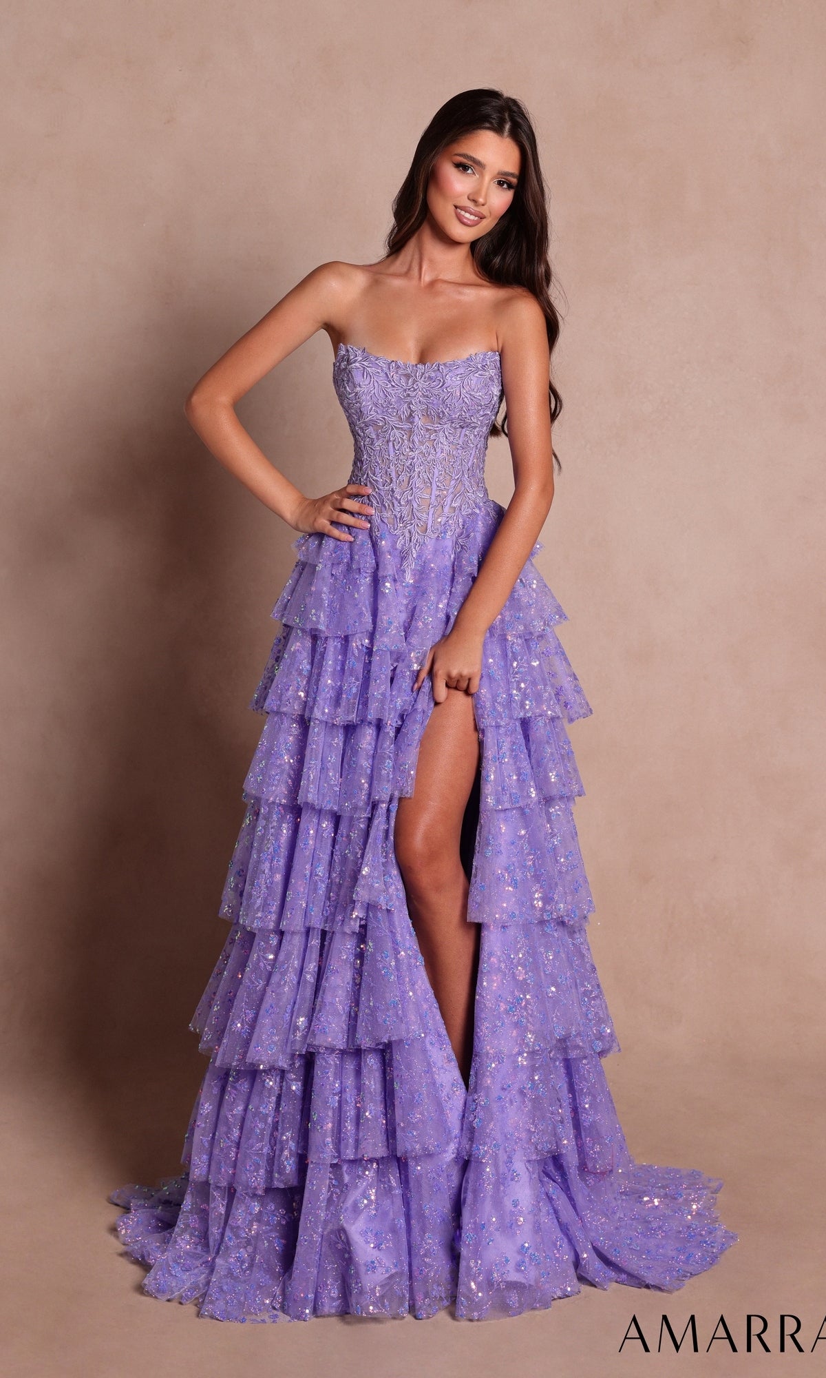 A woman is posing with one hand on her hip in front of a beige background, wearing the Amarra 89483 Strapless Ruffled Prom Ball Gown in lilac, which features tiered ruffles and sparkling sequins.