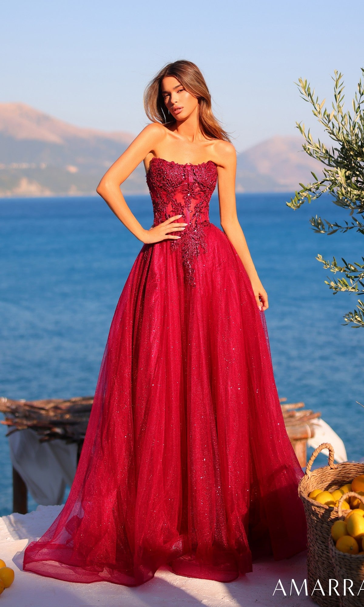 A woman in the Amarra 89486 long prom dress, a strapless glitter tulle gown with a lace-up back, stands outdoors by the sea with mountains behind her.