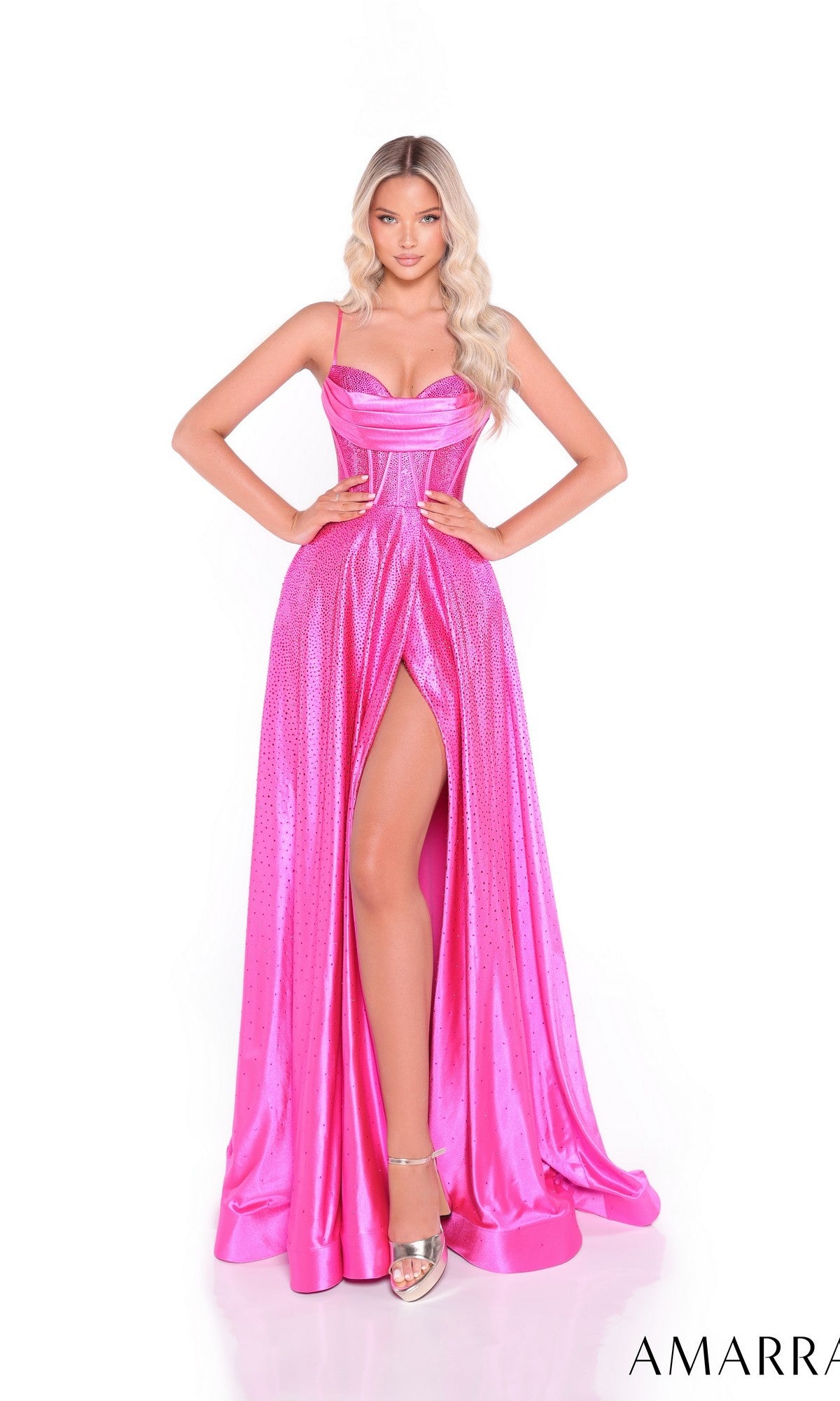 A woman models the Amarra 89522, a bright pink, floor-length satin prom dress with a high slit, spaghetti straps, and metallic sheen. She pairs it with silver heels and has long, wavy blonde hair.