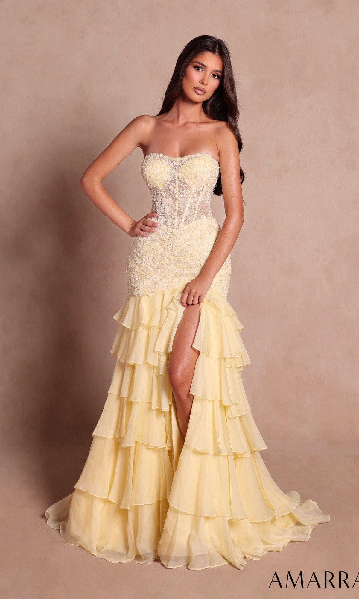 A woman wearing the Amarra 89631 long prom dress in light yellow, featuring a strapless design with a sheer bodice, floral details, and tiered ruffles along the skirt with a high slit, stands against a neutral background.