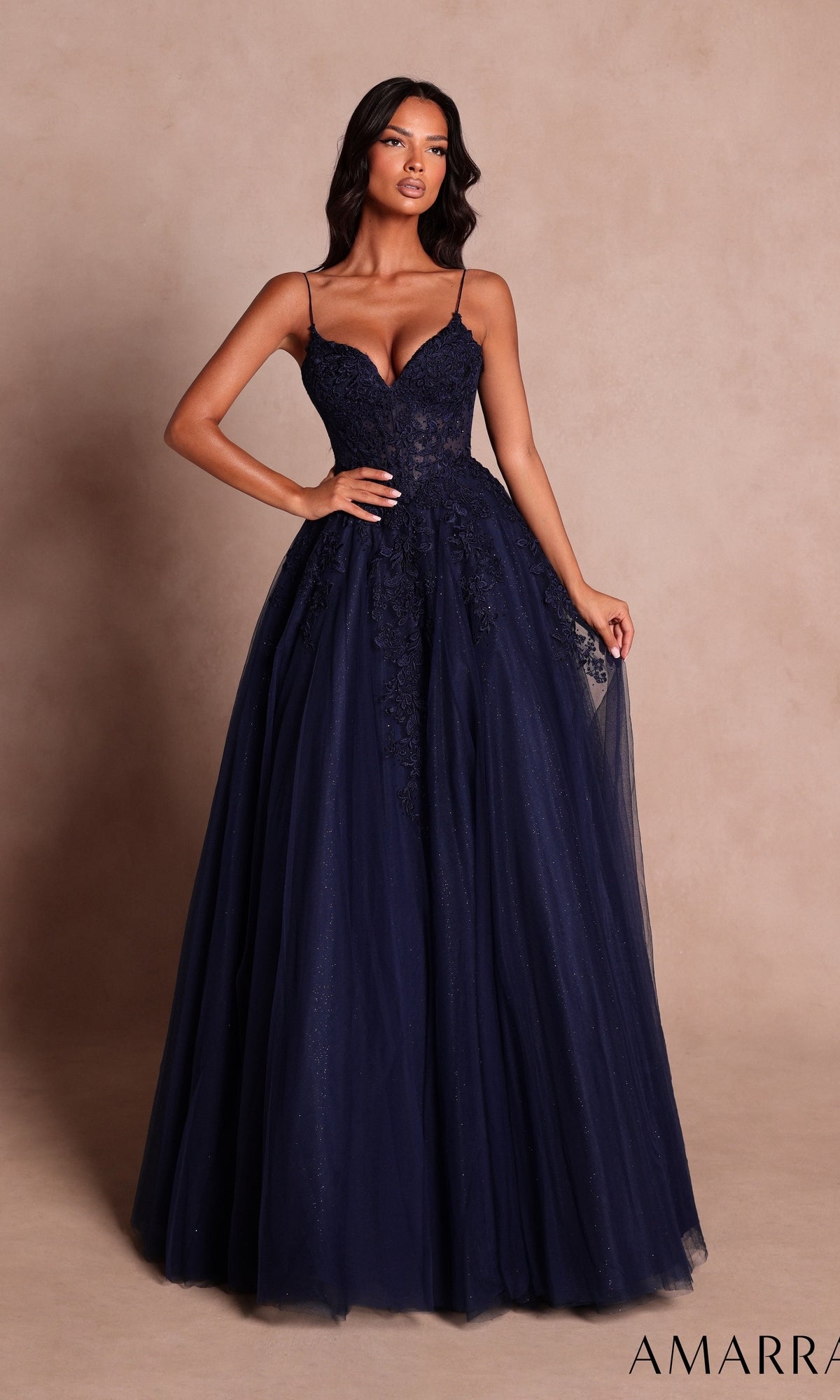 A woman wearing the Amarra 89682 long prom dress in navy, featuring a lace bodice with thin straps and a full tulle ball-gown skirt adorned with delicate floral appliqués, stands against a neutral background.
