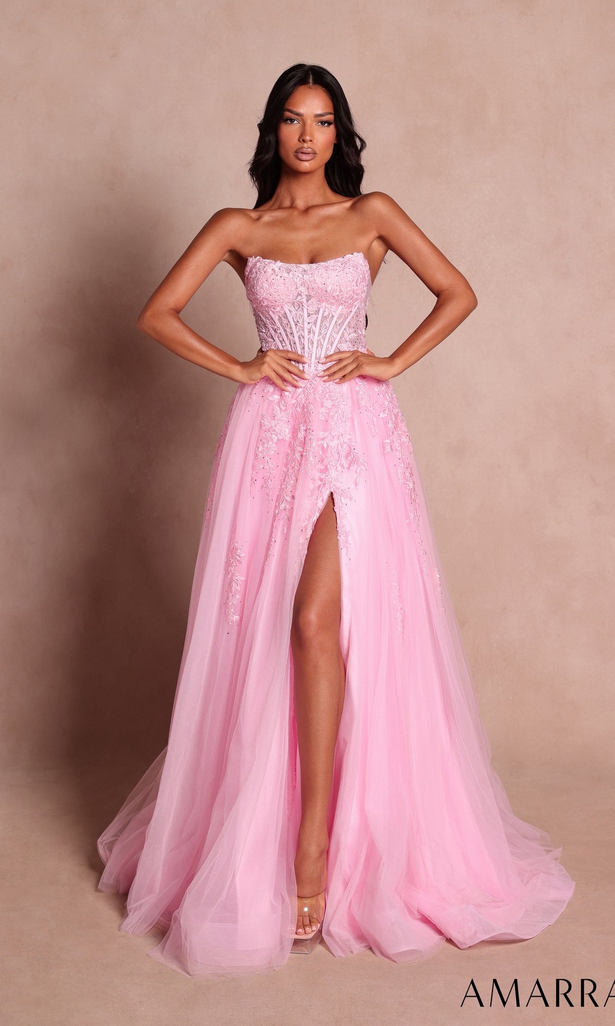 A woman stands against a beige background wearing the Amarra 89684 Long Prom Dress in light pink, featuring a strapless corset bodice, floor-length tulle A-line skirt, and a thigh-high slit.