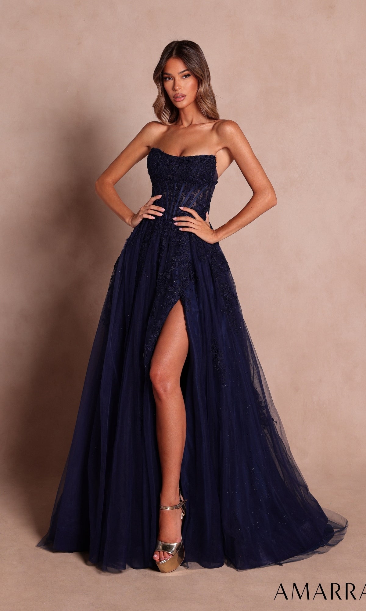 A woman wearing the Amarra 89684 long prom dress in navy, featuring a strapless neckline and a thigh-high slit, paired with metallic high heels, poses against a beige backdrop.