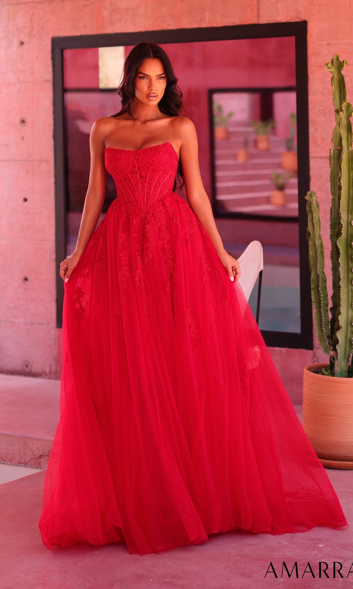A woman wearing the Amarra 89684 long red prom dress stands indoors beside a cactus, with a pink wall and large mirror in the background.