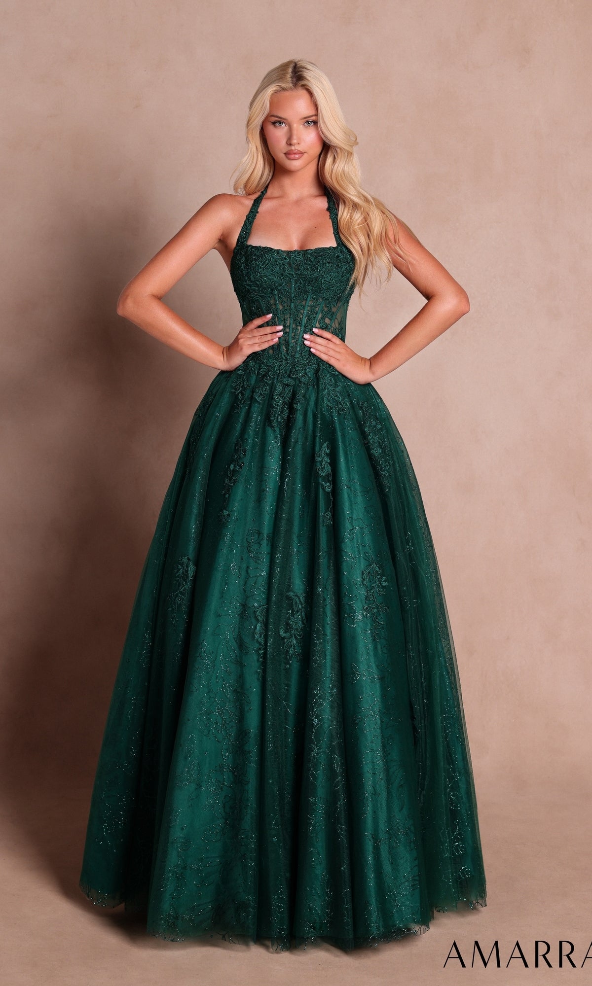 A woman stands against a plain beige background wearing the Amarra 89685 Prom Ball Gown in emerald—a floor-length dress with lace detailing, a glitter floral print, and a sheer corset bodice.