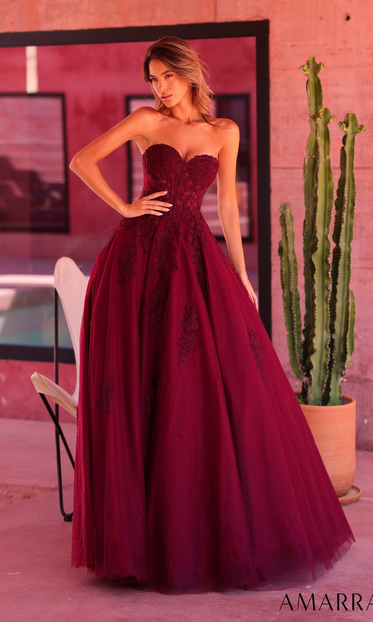 A woman wearing the Long Prom Dress: Amarra 89688 in plum, featuring a strapless neckline, delicate floral lace details, and a voluminous ball-gown skirt, stands indoors next to a tall potted cactus.