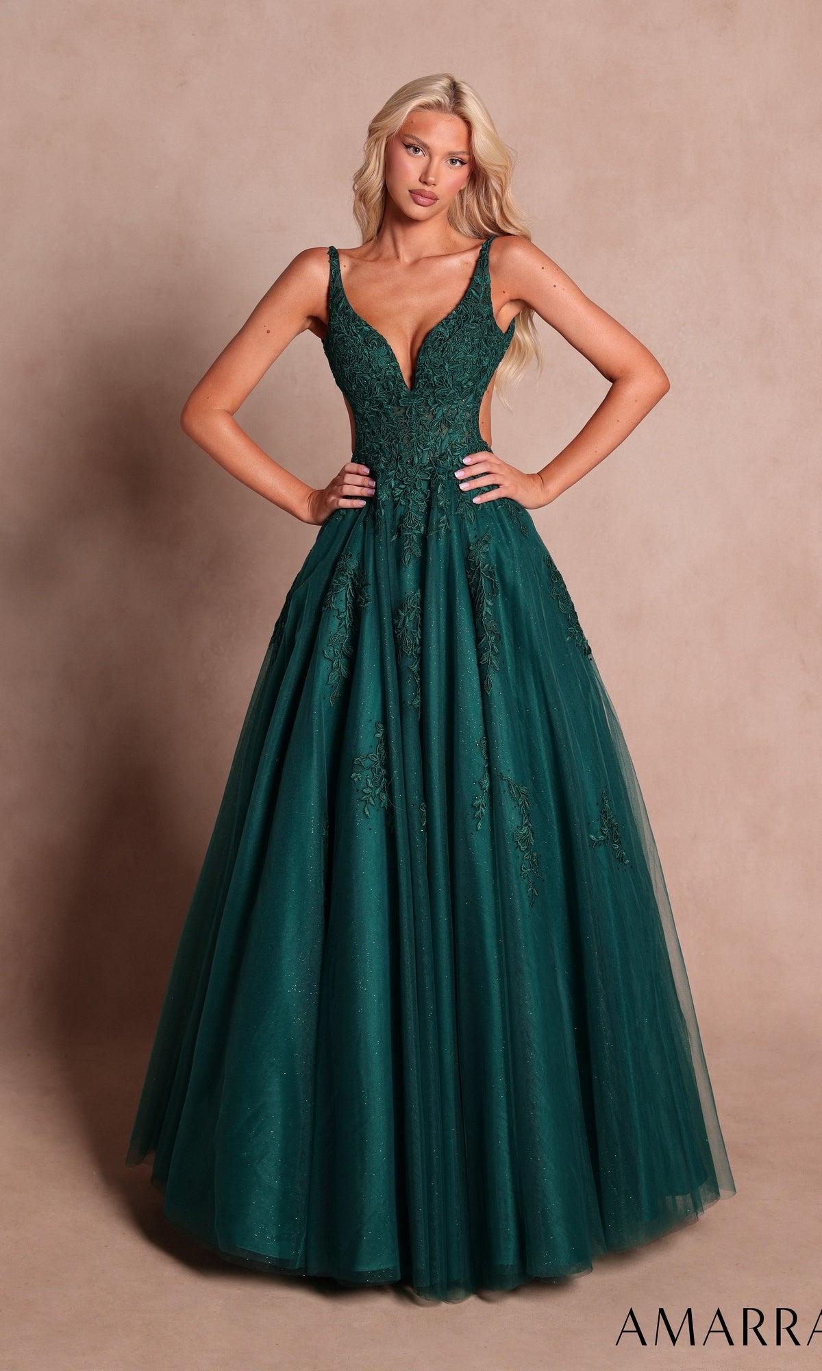 An elegant woman poses against a plain beige background wearing the Amarra 89786 long prom dress in emerald—a sleeveless gown featuring a fitted bodice embellished with floral lace appliqués and a flowing tulle skirt.