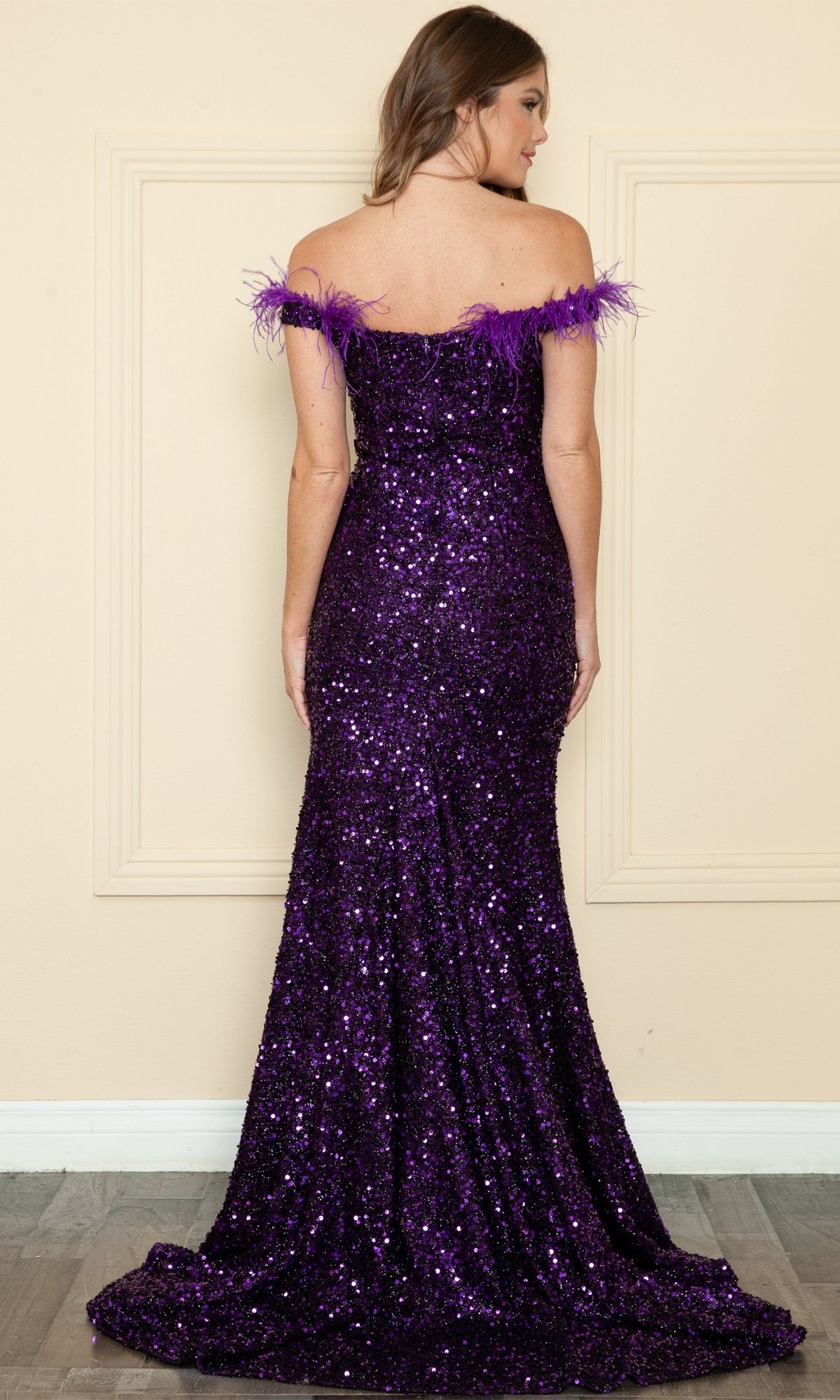 A woman stands facing away from the camera, wearing the Sequin-Velvet Long Feathered Prom Dress 8980—an off-the-shoulder purple gown with sequin details and feather trim—set against a beige wall.