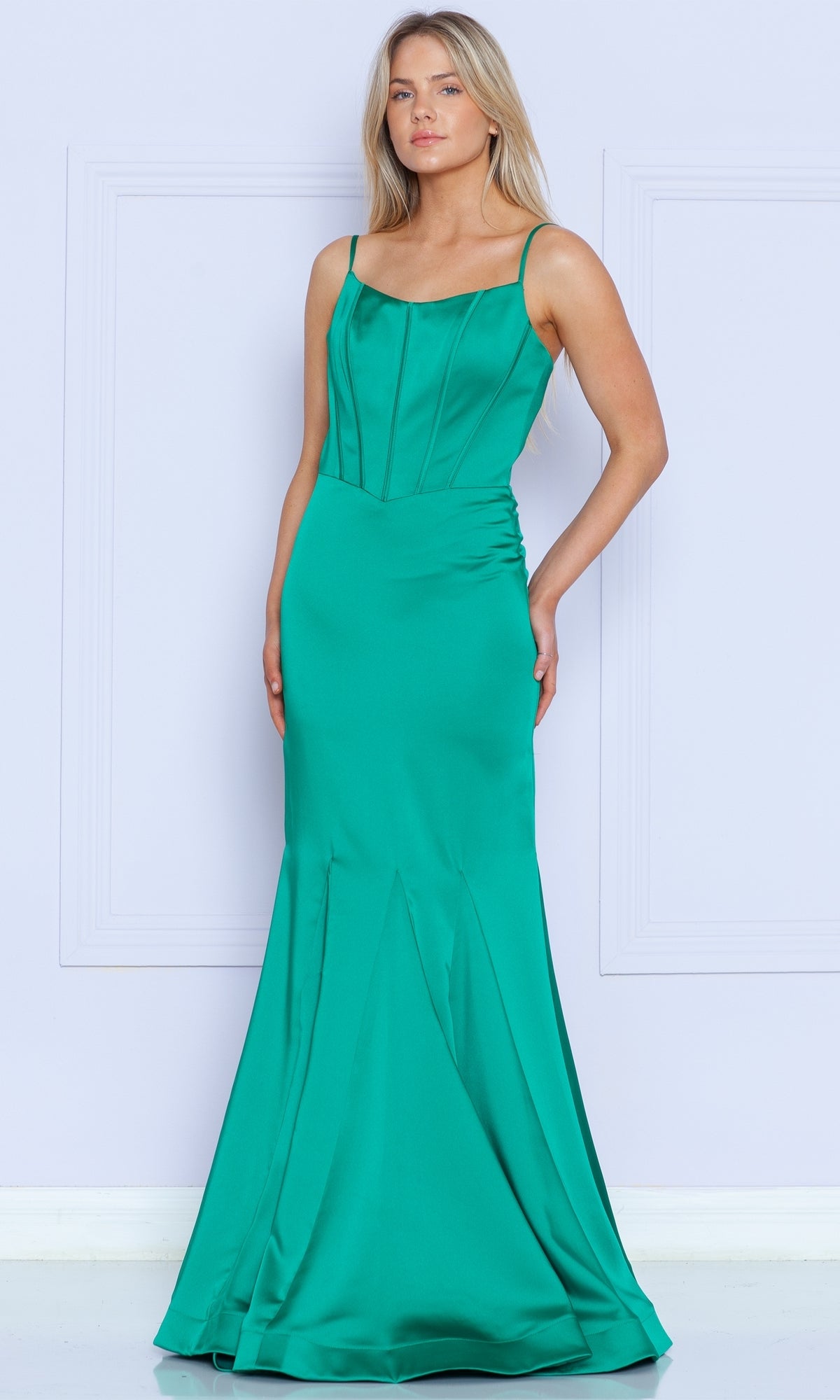 A woman poses against a light wall in the Stretch-Charmeuse Long Mermaid Prom Dress 9006, an emerald-colored, sleeveless gown with thin straps and a fitted, flared floor-length skirt.
