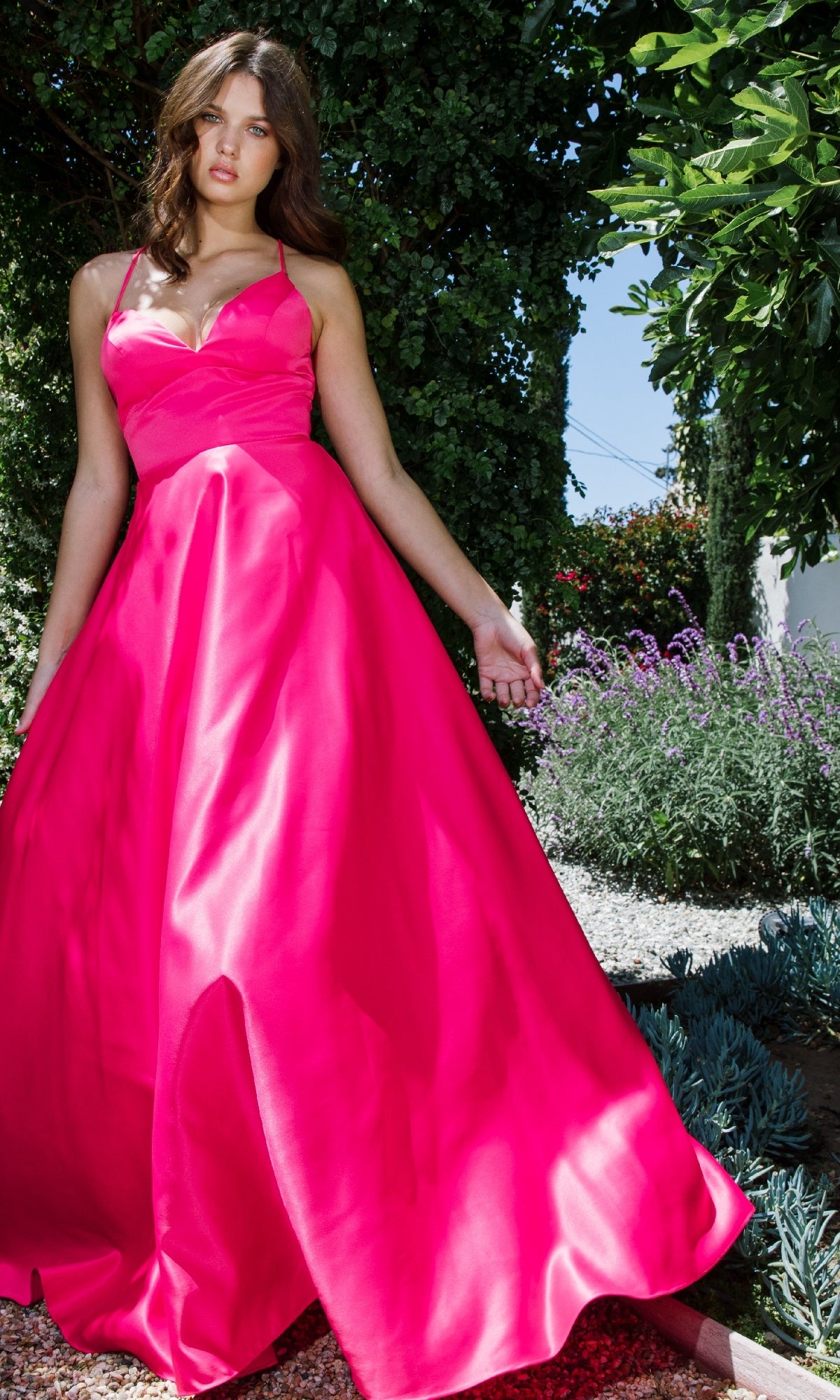 A woman stands outdoors in a garden wearing the Eureka 9008 Strappy-Back Long Satin Prom Dress, featuring a bright pink hue, thin straps, and a fitted bodice.