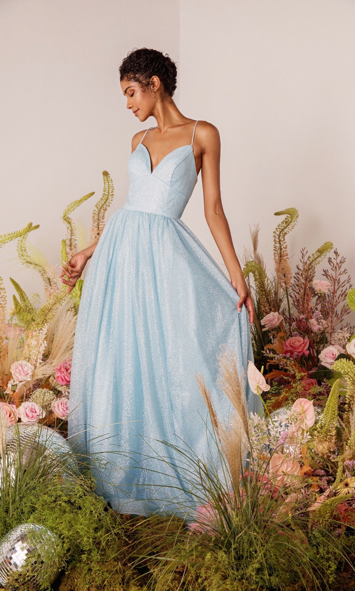 A woman in the Strappy-Back Glitter Prom Ball Gown: Eureka 9009, a light blue a-line dress with spaghetti straps, stands among flowers and greenery, holding one side of her gown.