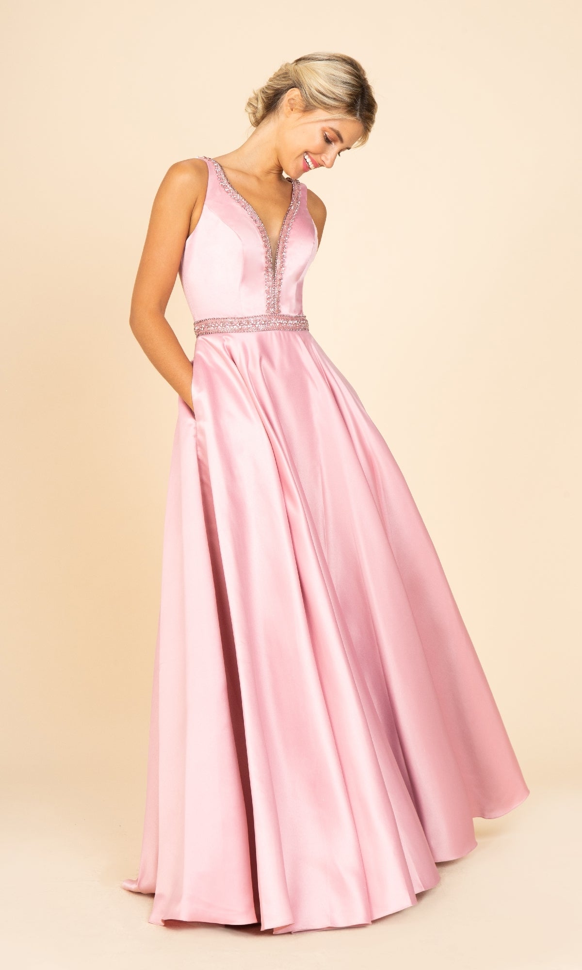A woman in the Beaded-Waist Long Satin Prom Dress (Eureka 9010)—a sleeveless, floor-length pink gown with a V-neckline and beaded waist—stands against a beige background, looking down with her hands in her pockets.