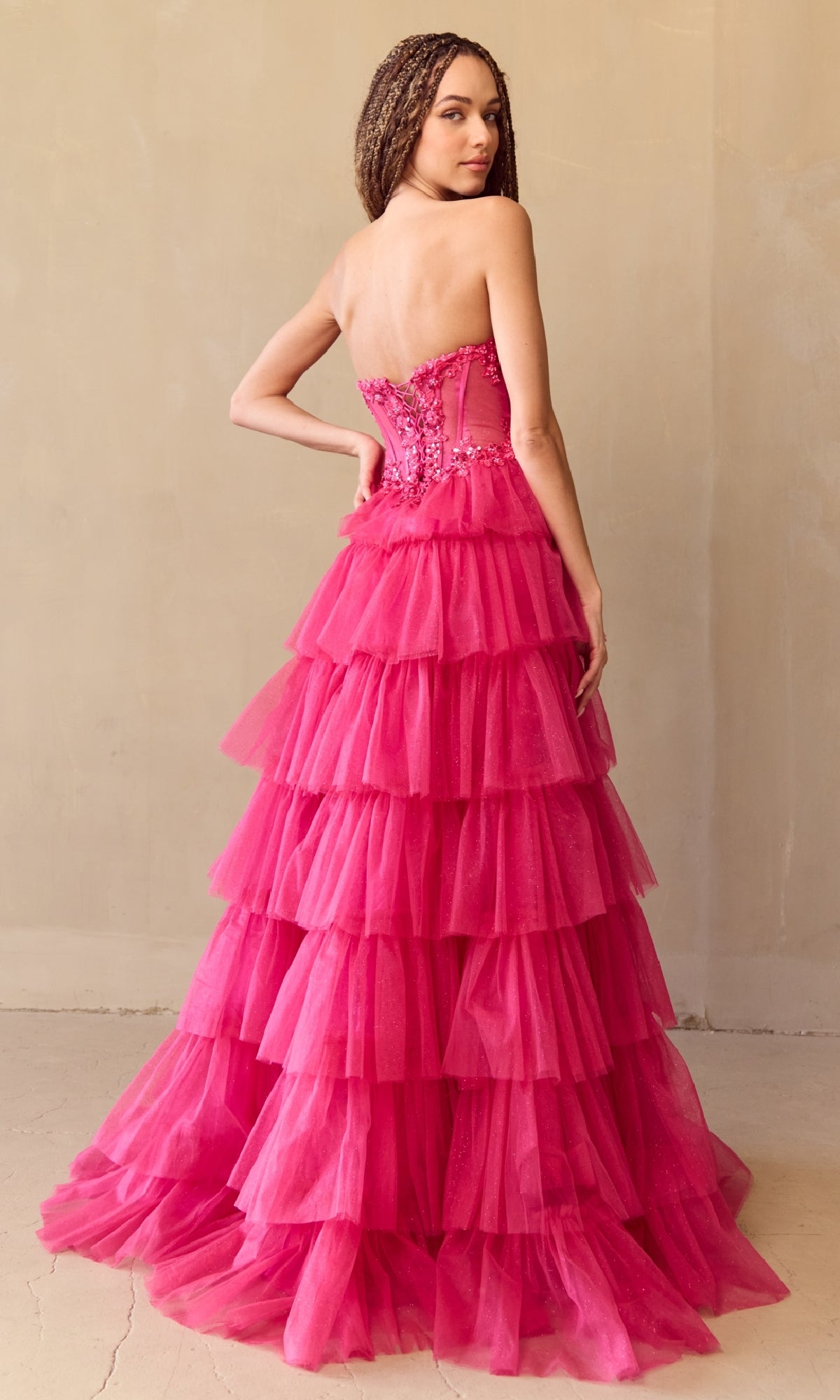 Model wearing the Amelia Couture 9053 Strapless Long Prom Ball Gown, featuring a beaded corset bodice and layered hot pink tulle skirt, poses with one hand on her hip against a plain beige background.
