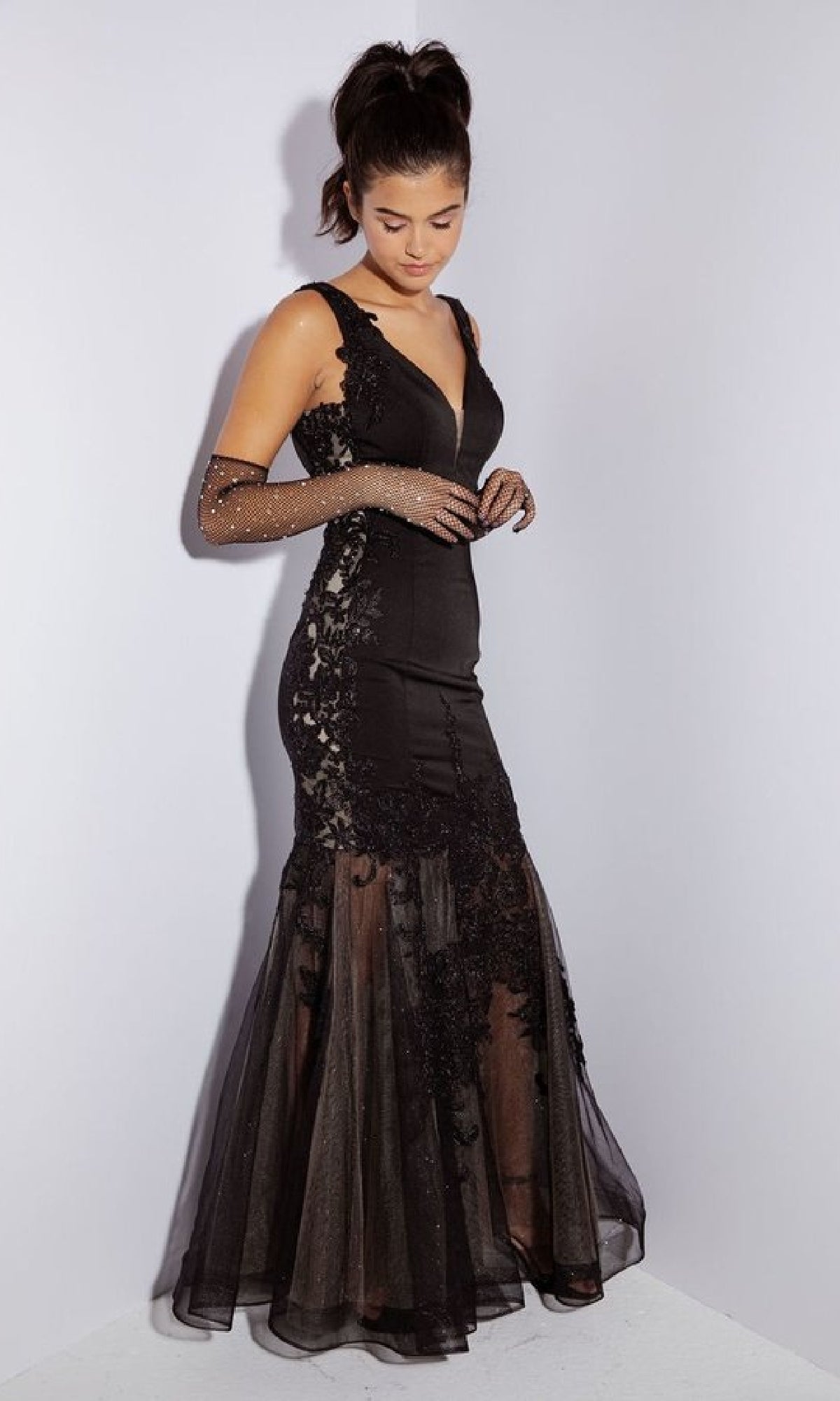 A woman stands against a white wall wearing the Eureka 9060 long black mermaid prom dress with lace and sheer details, paired with matching sheer gloves.