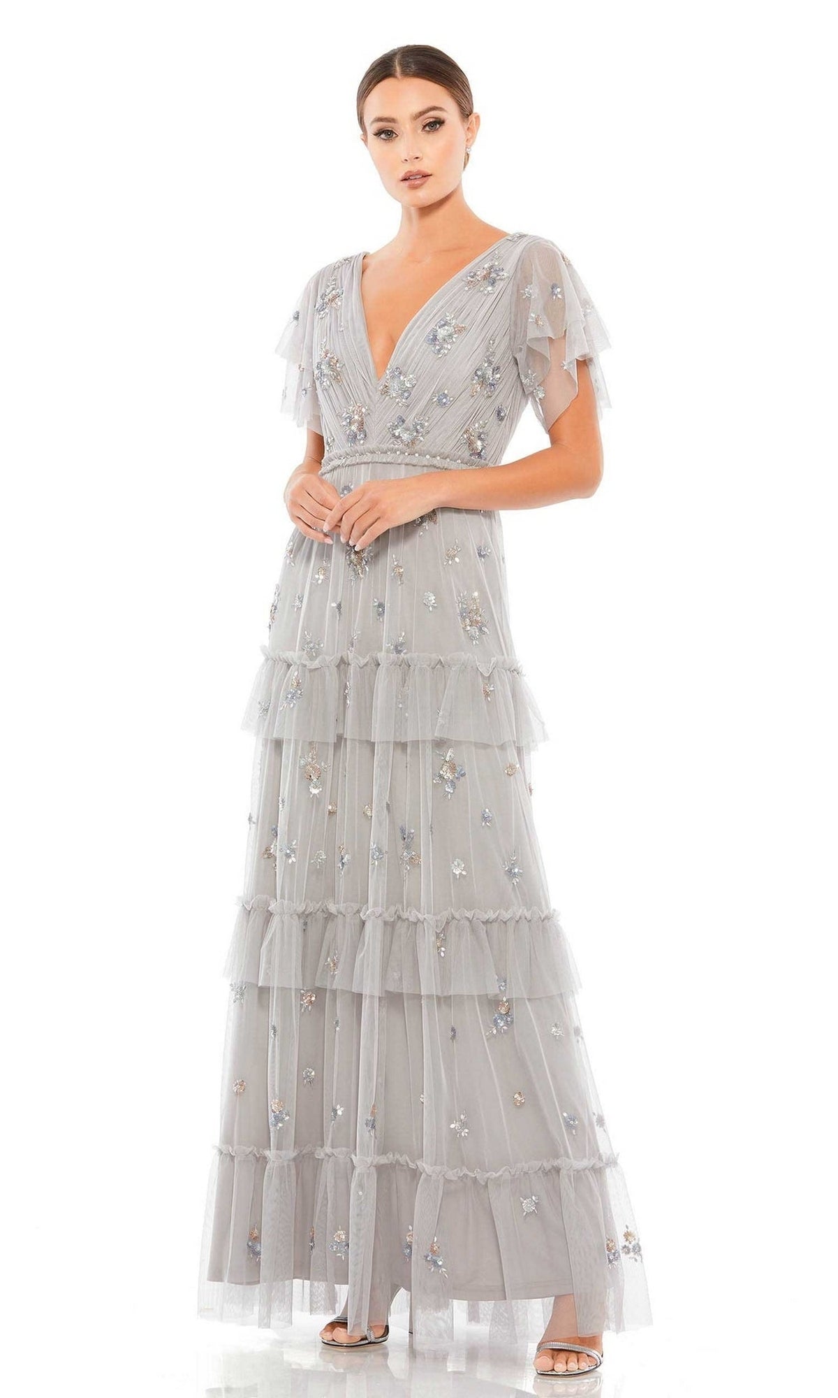 A woman stands wearing the Mac Duggal 9068 Short Sleeve Platinum Formal Gown, its floral-embellished, floor-length tiered tulle design shining elegantly against a white background.