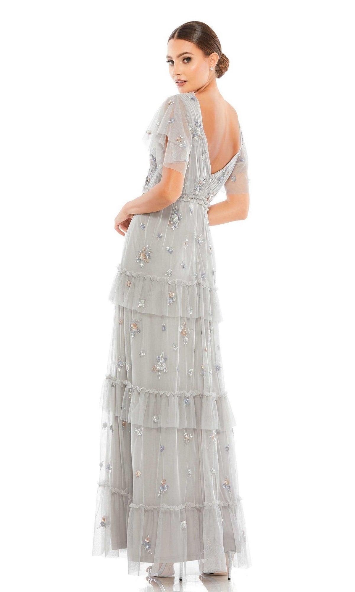 A woman in the Short Sleeve Platinum Formal Gown: Mac Duggal 9068, adorned with floral embellishments, stands facing away and looks over her shoulder in her elegant beaded evening dress.