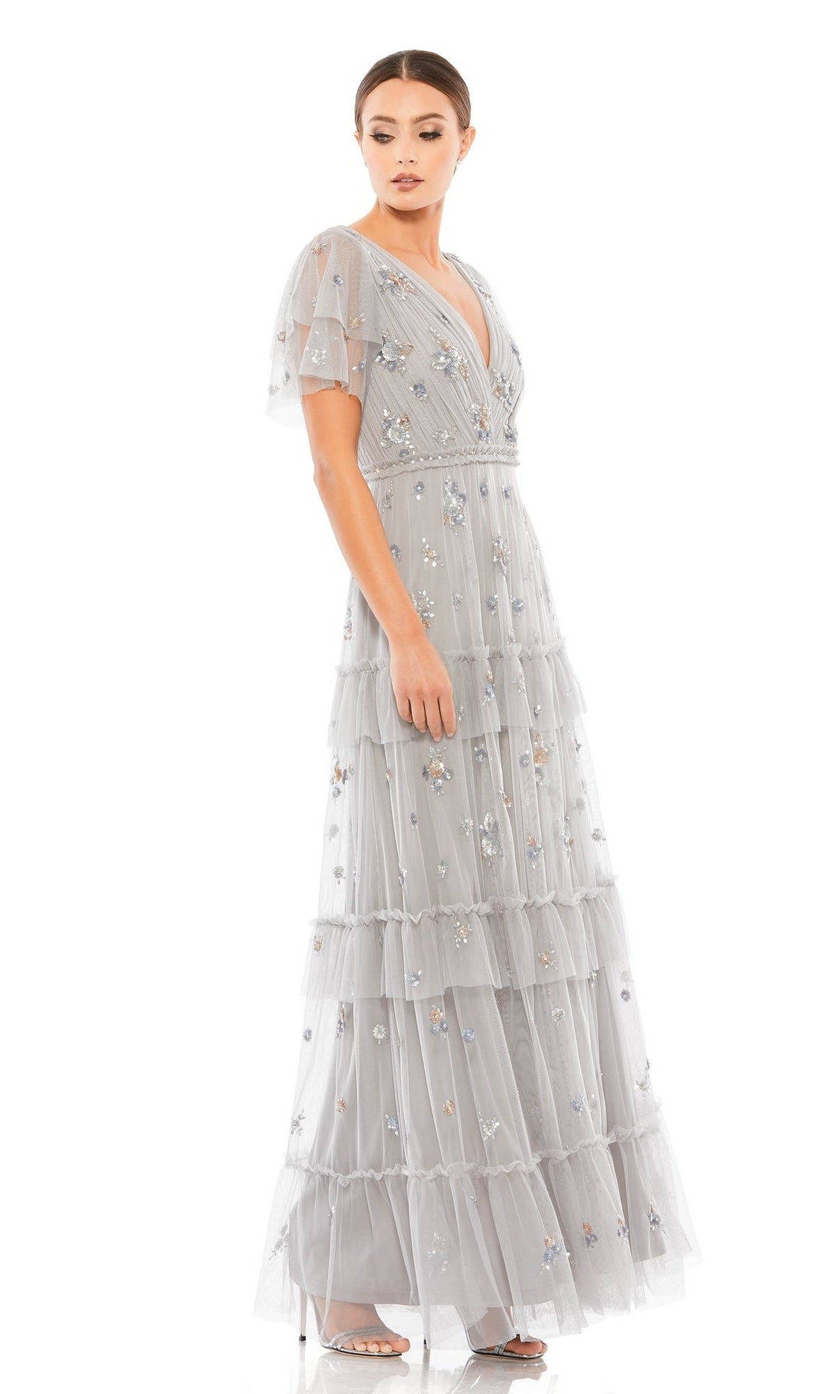 A woman models the Mac Duggal 9068 Short Sleeve Platinum Formal Gown, a light grey, floor-length dress with tiered layers and floral embellishments, facing slightly sideways against a white background.