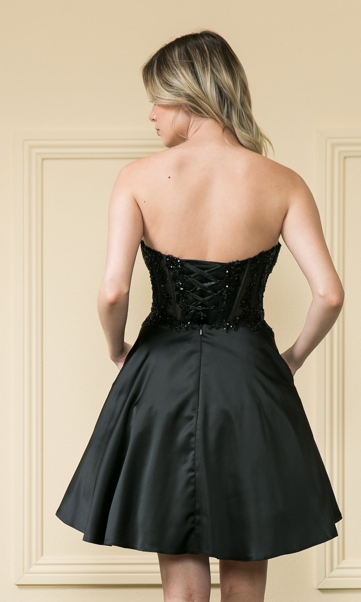 A woman faces away wearing the Corset-Style Strapless Short Homecoming Dress 9084, featuring a lace-up back and flared skirt.