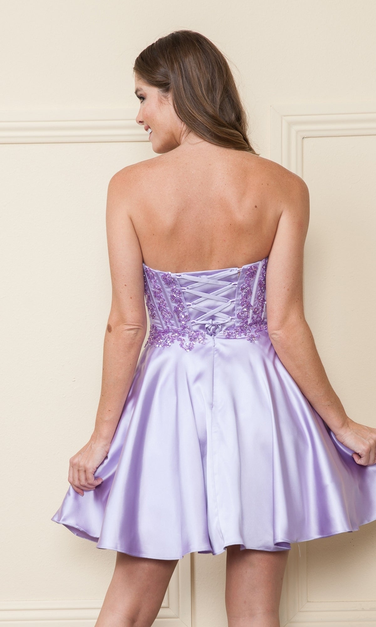 A woman faces away, gently holding out the lavender satin skirt of the Corset-Style Strapless Short Homecoming Dress 9084, highlighting its beaded corset bodice, embroidered details, and lace-up back.
