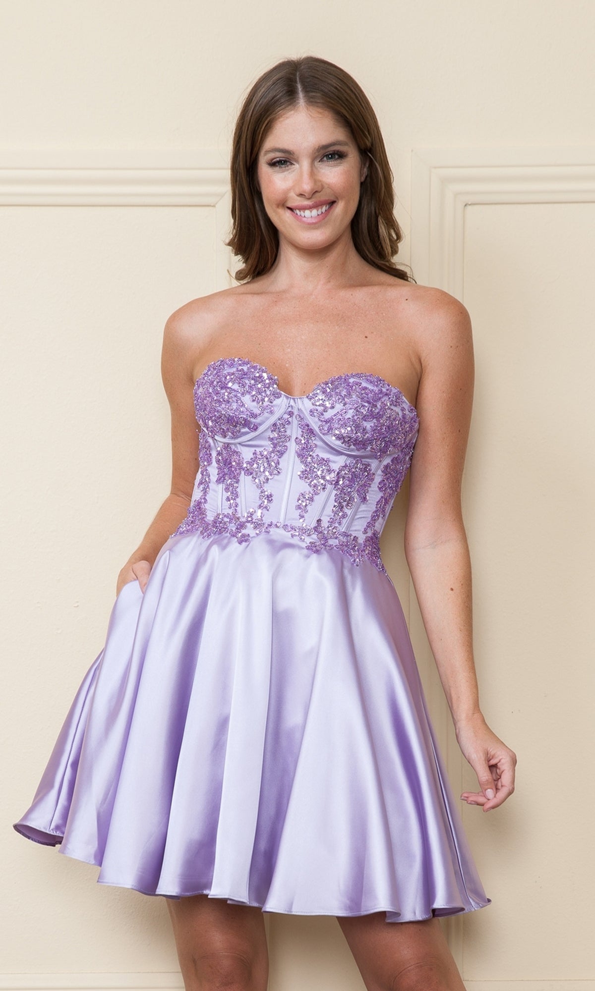 A woman smiles against a cream wall, wearing the Corset-Style Strapless Short Homecoming Dress 9084 in lavender satin, featuring a beaded corset bodice and short silhouette.
