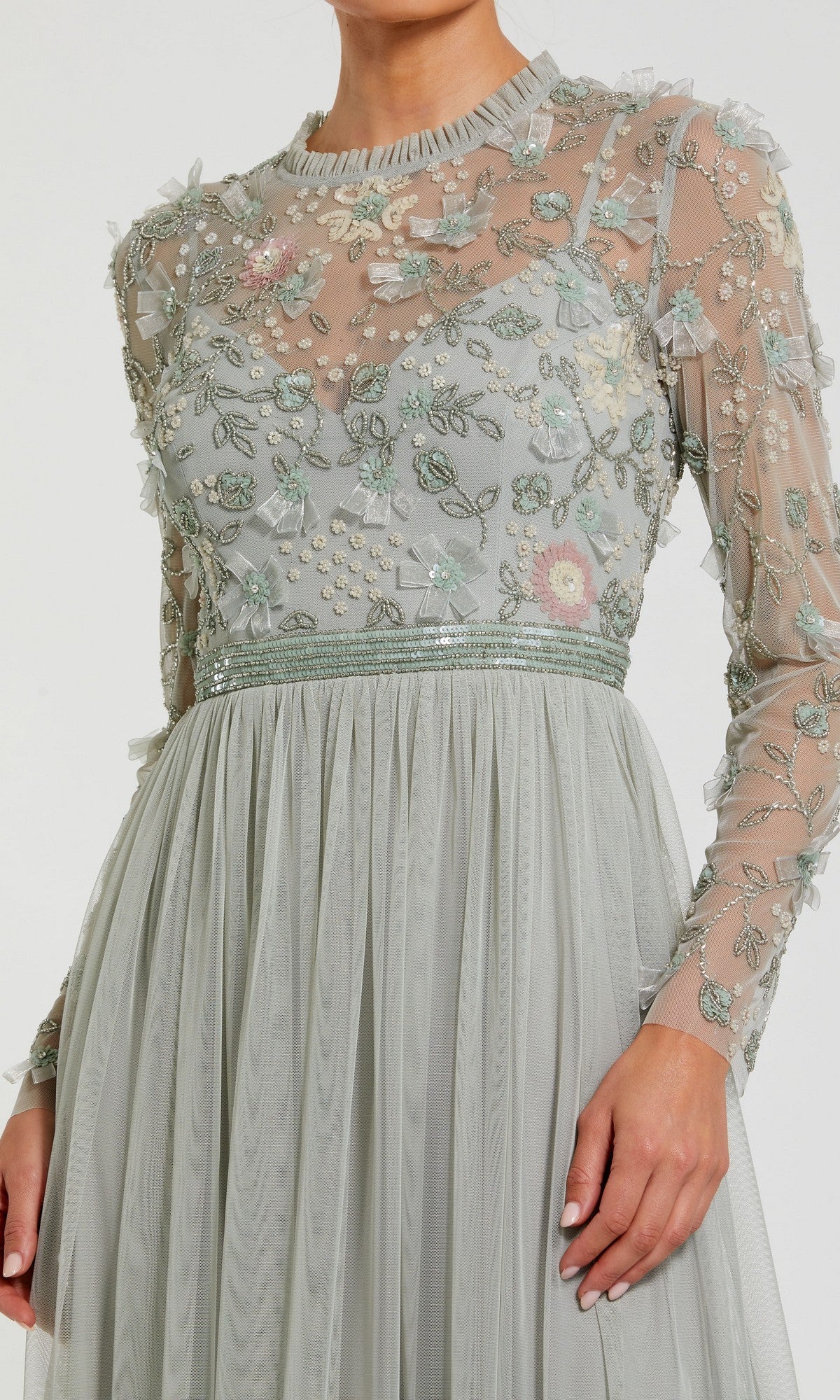 A woman wears the Mac Duggal 9132 Long Sleeve Vanilla Formal Gown featuring sheer long sleeves, a floral embroidered and beaded bodice, and a pleated A-line skirt.