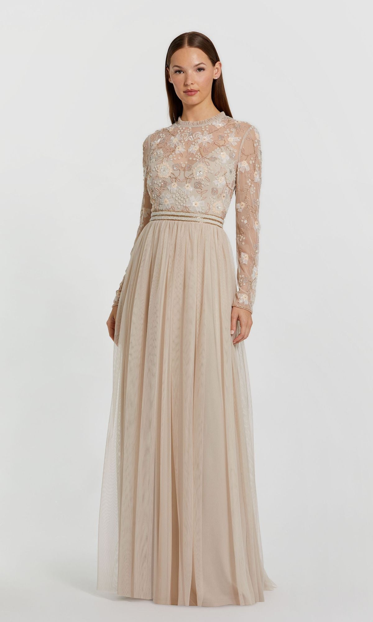 A woman stands against a white background wearing the Mac Duggal 9132 Long Sleeve Vanilla Formal Gown, featuring sheer embroidered sleeves and a fitted, floral-embroidered bodice in a chic beige hue.