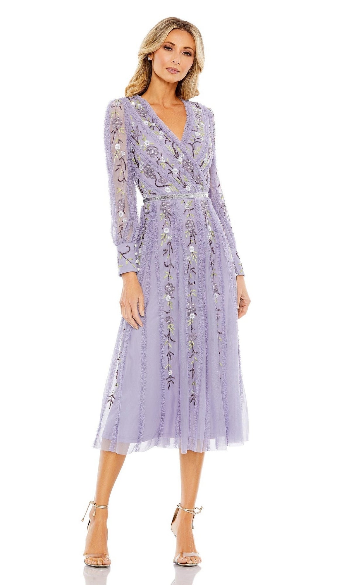 A woman stands in heels against a white background, wearing the Knee Length Wedding Guest Dress 9133 by Mac Duggal—a long-sleeve, lavender A-line midi dress featuring floral embroidery and a sheer overlay.