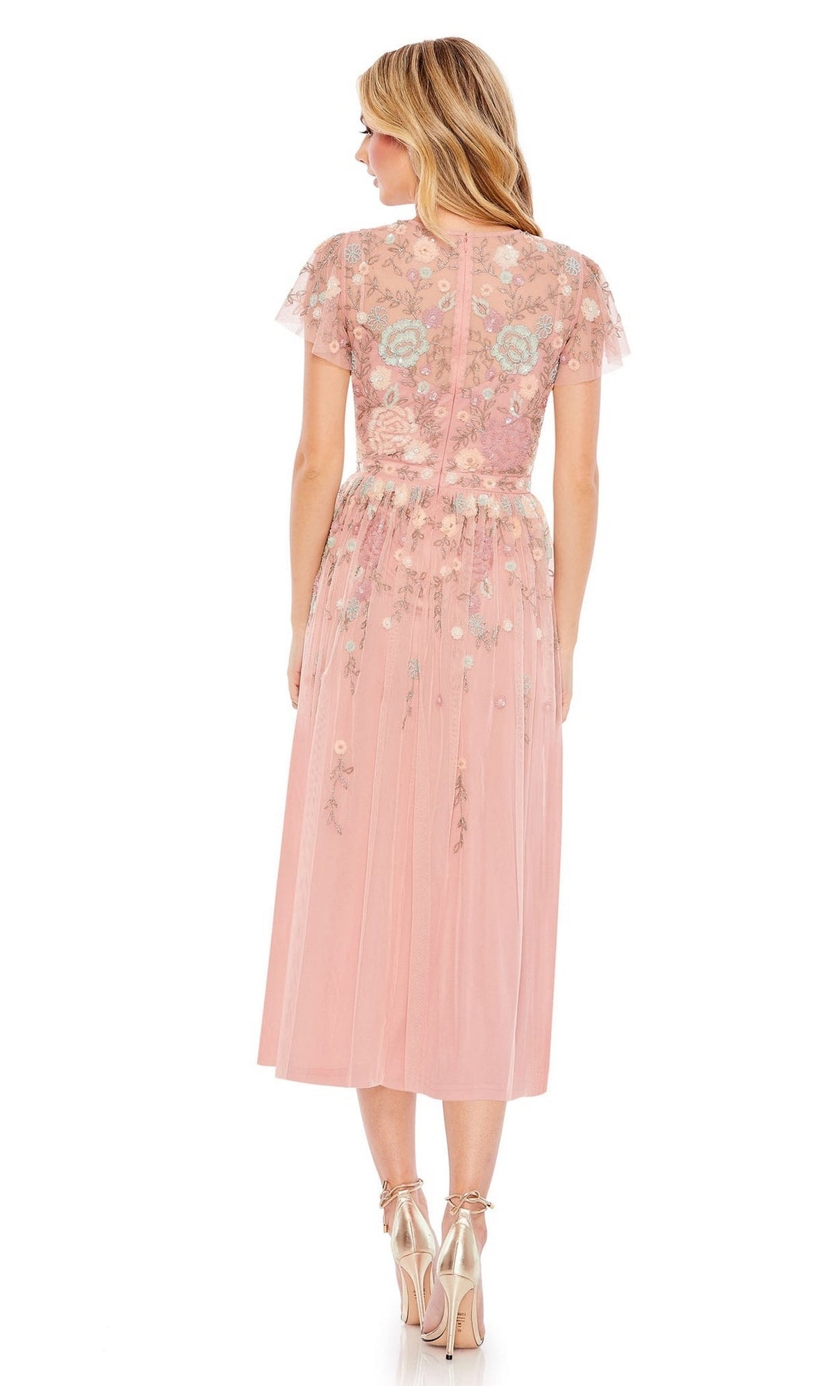 A woman stands facing away, wearing the Knee Length Wedding Guest Dress 9134 by Mac Duggal—a pink floral beaded midi A-line dress with short sleeves—paired with metallic high-heeled shoes.