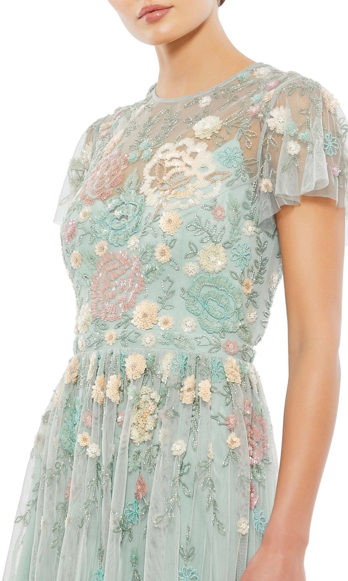 A woman wears the Knee Length Wedding Guest Dress 9134 by Mac Duggal—a sheer, beaded floral dress with pastel embroidery over a light green base, perfect for a chic wedding guest look.