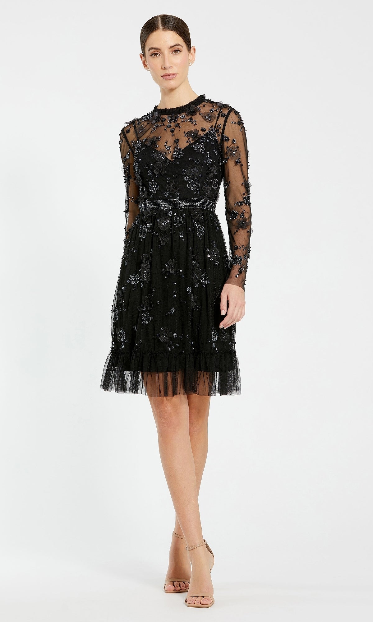 A woman wears the Mac Duggal 9140 Short Black Wedding Guest Dress with sheer long sleeves and floral embroidery, posing with one leg forward against a plain background—a chic option for any wedding guest.