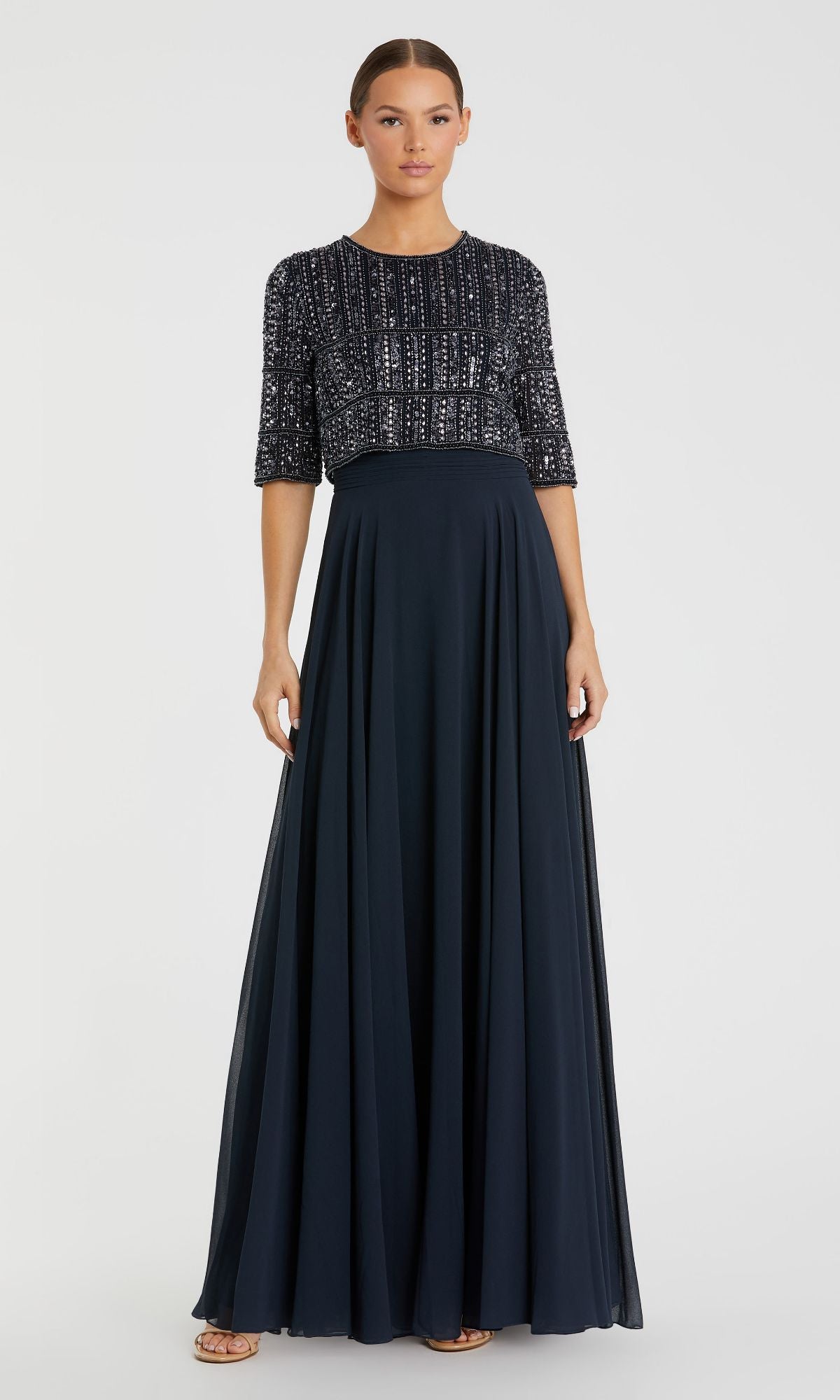 A woman stands facing forward in the Mac Duggal 9180: a navy, floor-length formal dress featuring short sleeves, a beaded bodice, and a flowing A-line skirt.