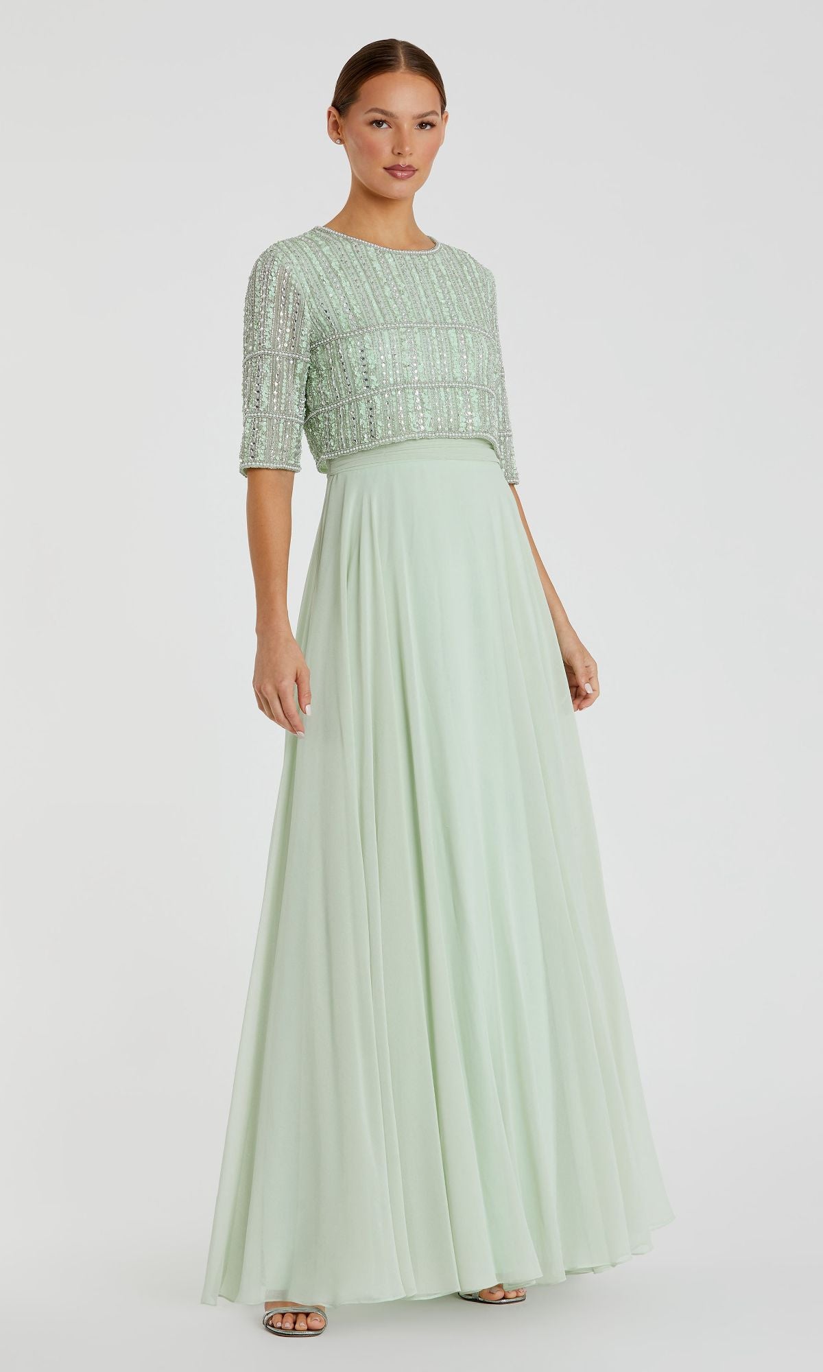 A woman wears the Mac Duggal 9180—a short sleeve, long formal dress with a sequined, beaded crop top and flowing skirt—in light green, standing against a plain white background.