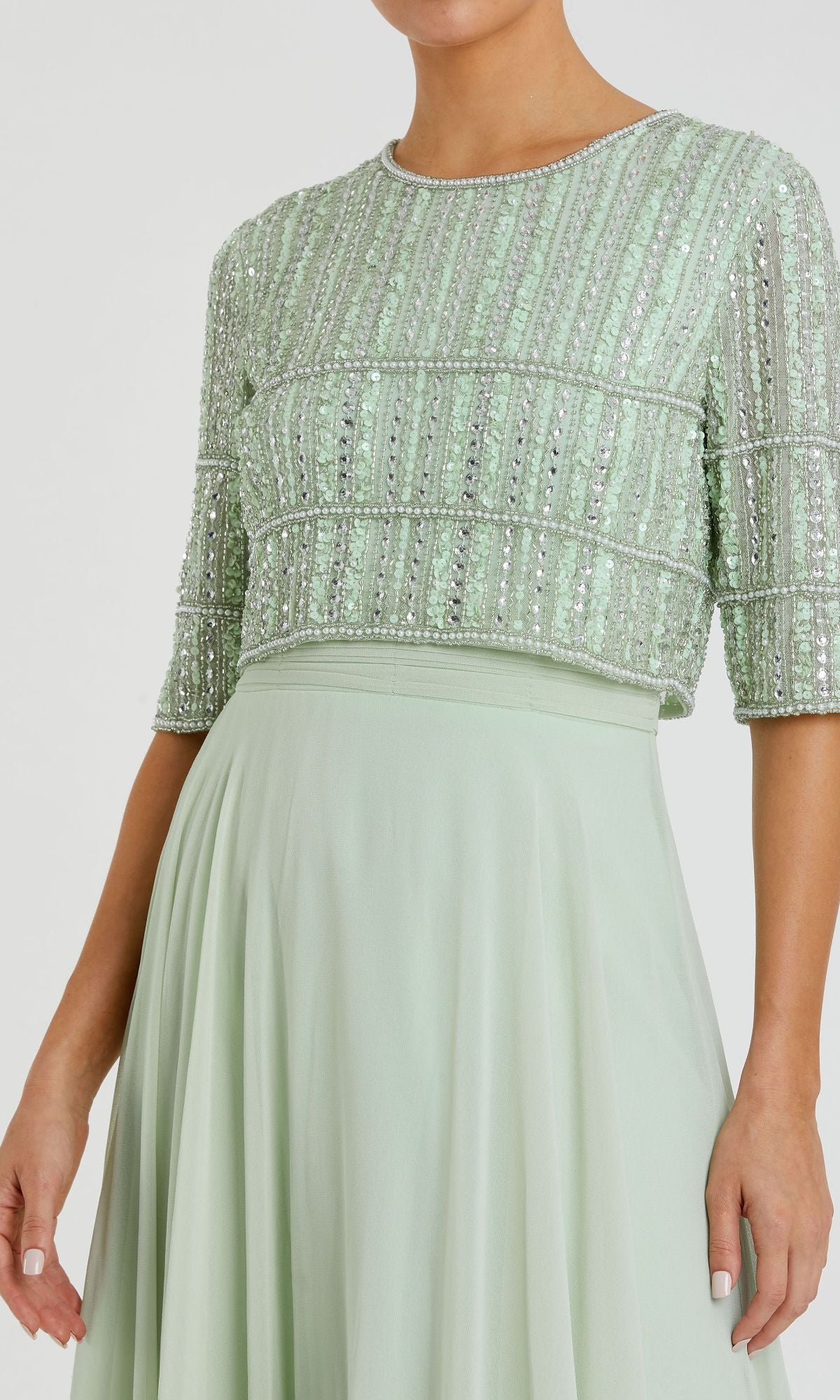 A woman wearing the Mac Duggal 9180: a short sleeve long formal dress with a sequined beaded crop top and a flowing light green A-line skirt.