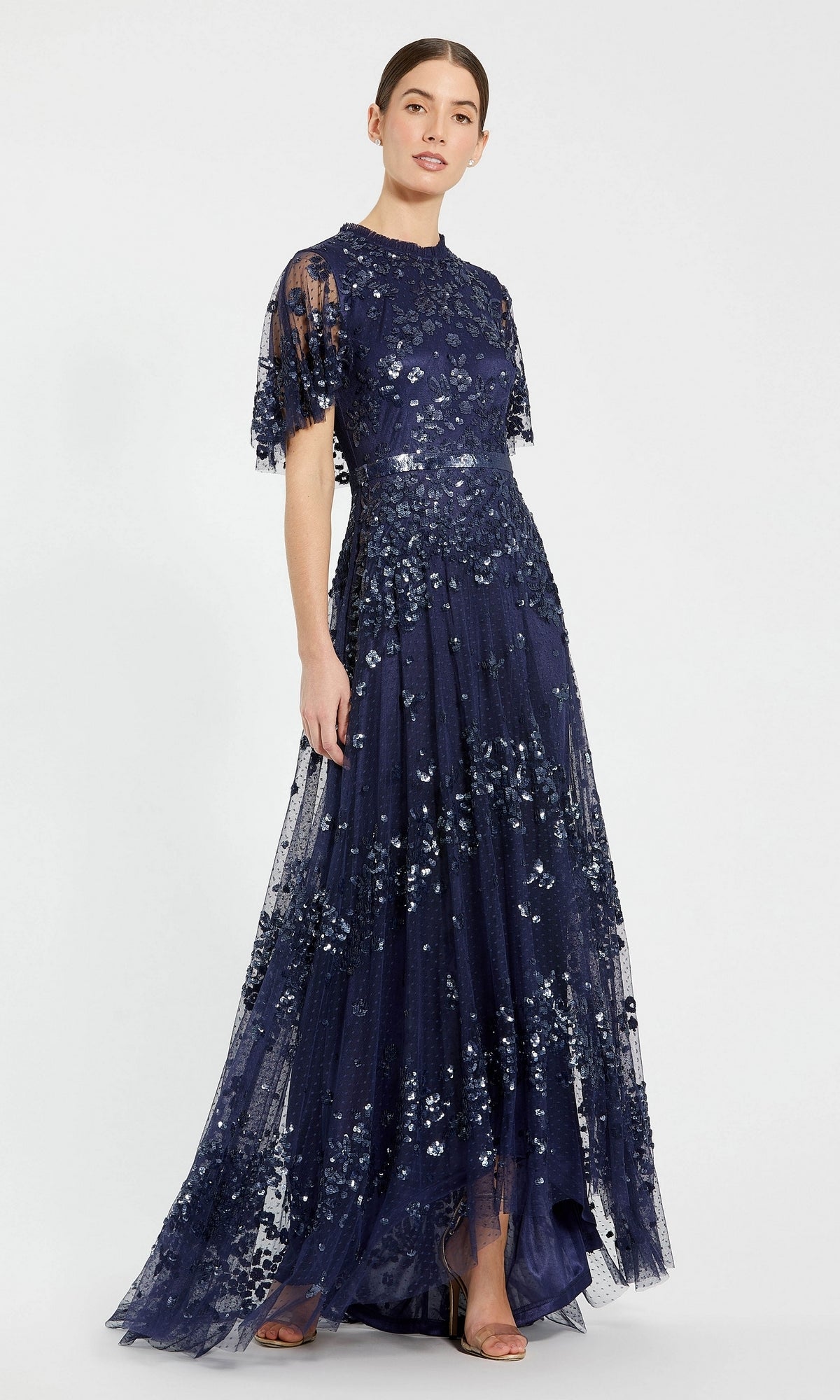 A woman stands against a plain background wearing the Mac Duggal 9192 Short-Sleeve Blue Formal Dress, featuring a floor-length design with sequins, floral embellishments, and a sheer overlay.