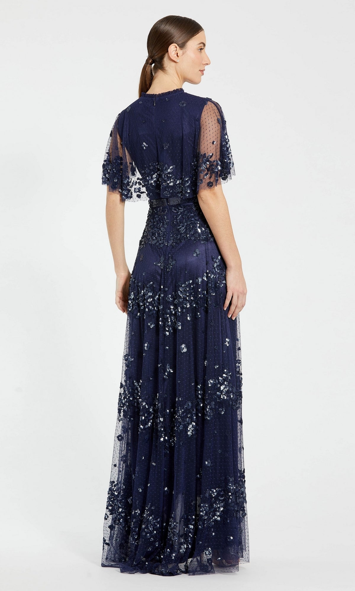 A woman stands with her back to the camera, wearing the Mac Duggal 9192 short-sleeve blue formal dress, a floor-length evening gown featuring elegant floral embellishments.