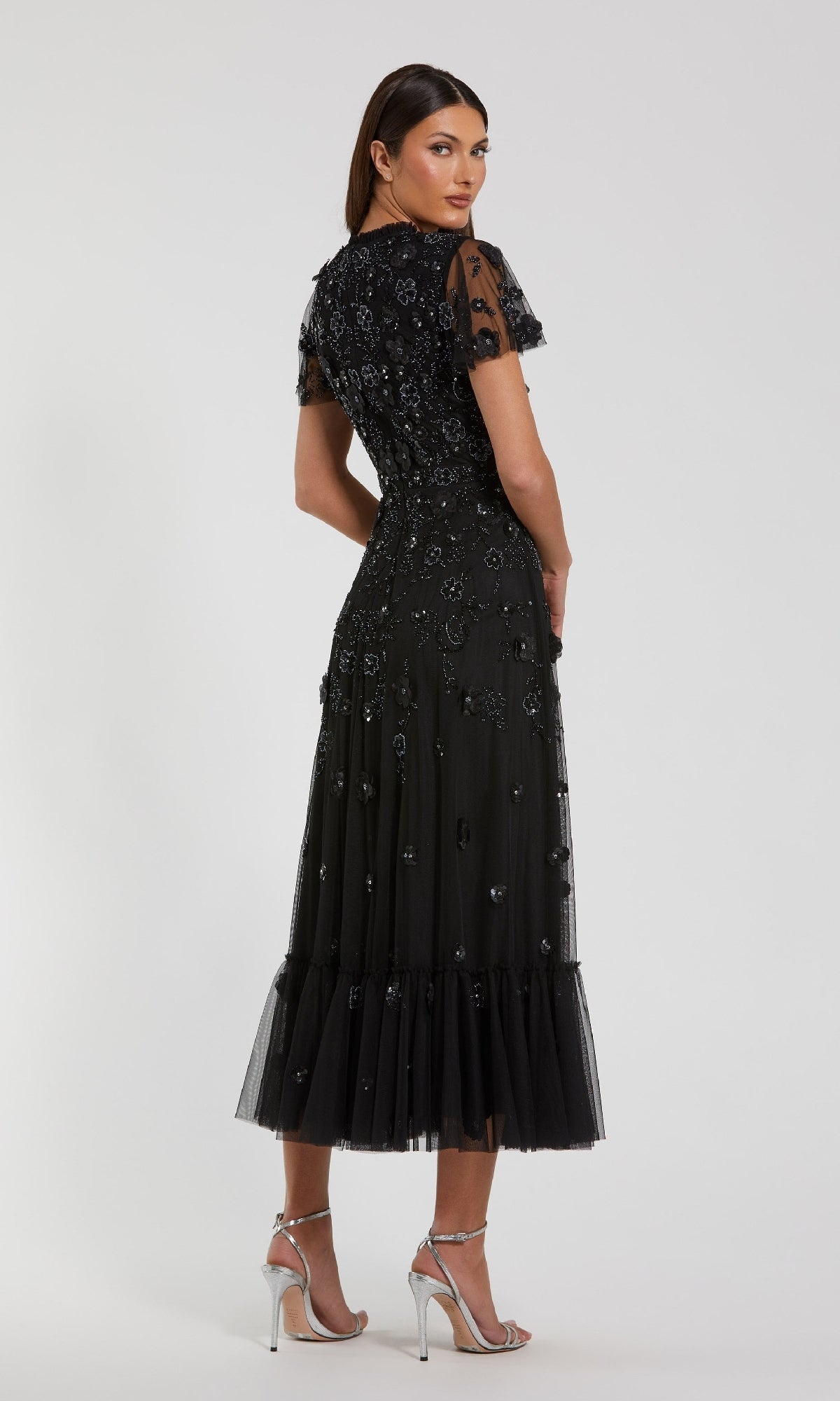 A woman stands facing away from the camera in a Mac Duggal 9212 Floral Midi Wedding-Guest Dress with short sleeves, ruffle hem, and silver high-heeled sandals against a plain background.