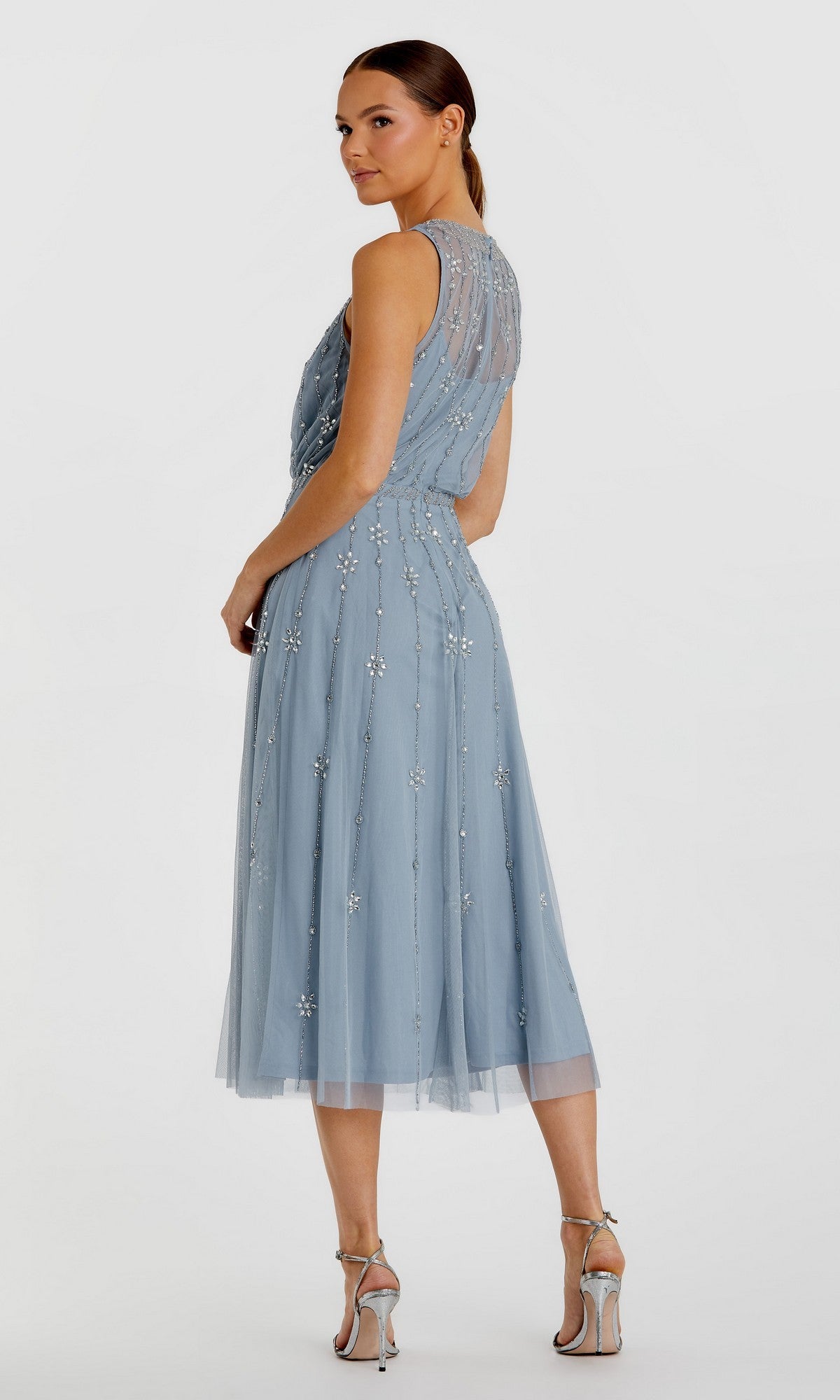 Woman in the Mac Duggal 9222 Midi Wedding Guest Dress—sleeveless, light blue with beaded floral details and a knee-length A-line silhouette—poses against a plain backdrop, paired with silver high-heeled sandals.