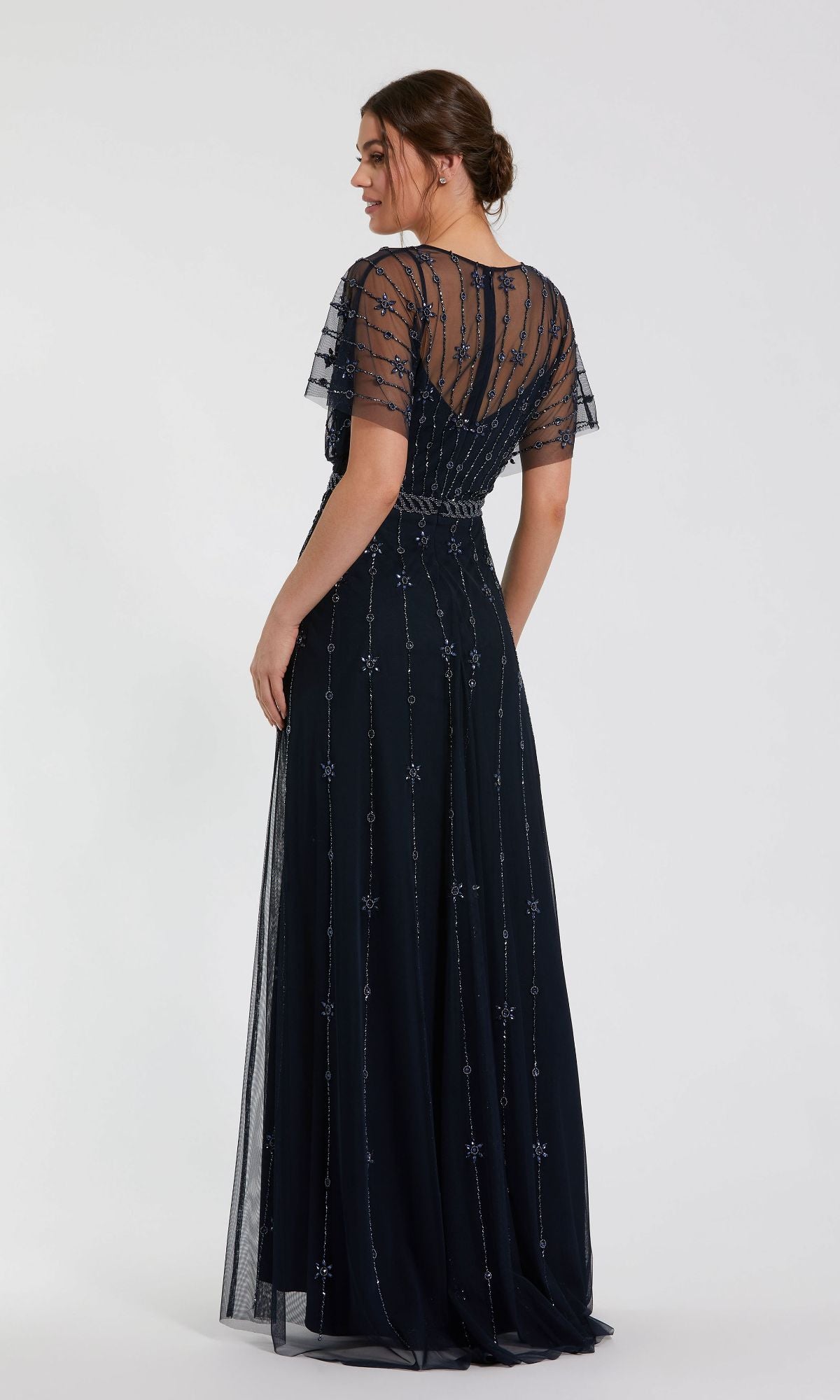 A woman wearing the Mac Duggal 9223 Short Sleeve Long Formal Dress with sheer short sleeves, intricate beadwork, and a fitted waist stands facing away from the camera.