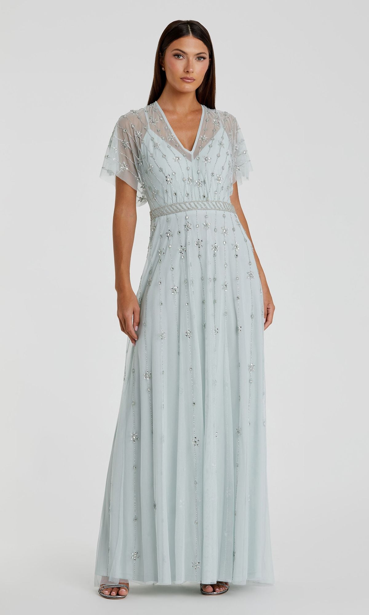 A woman stands against a plain background wearing the Mac Duggal 9223, a short sleeve long formal dress in light blue with sheer sleeves and delicate silver embellishments.