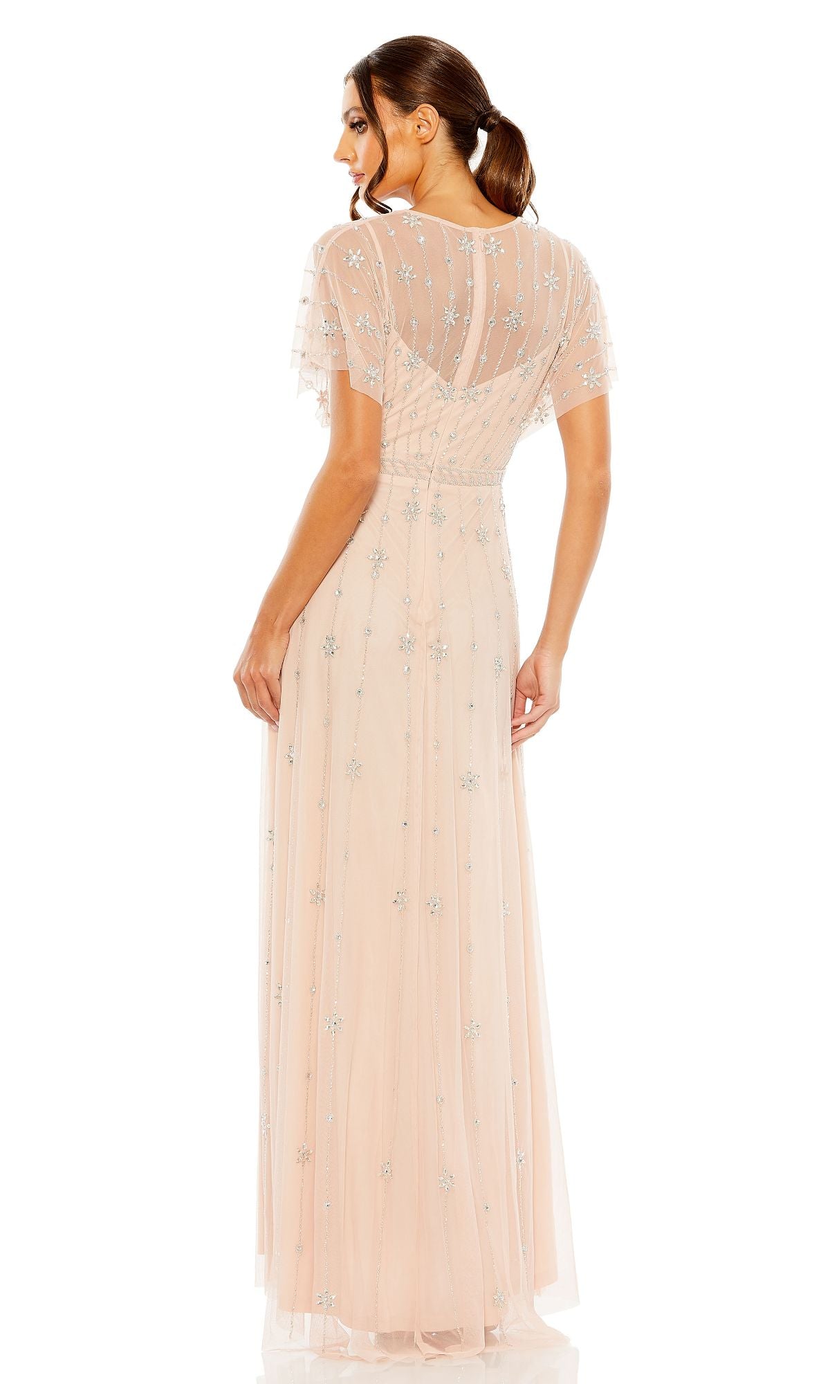 A woman stands facing away, wearing the Mac Duggal 9223 short sleeve long formal dress in light pink with silver floral embellishments and sheer sleeves.