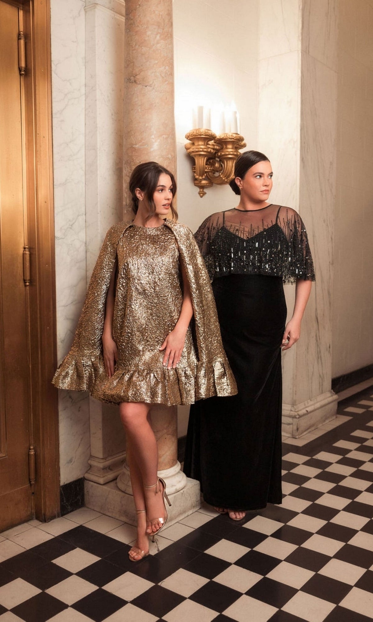Two women stand indoors on a black-and-white checkered floor; one wears an embellished sheer cape dress, while the other wears the Long Plus-Size Formal Dress: Mac Duggal 9228, a black gown with beaded detail. Marble columns and a wall sconce complete the elegant scene.