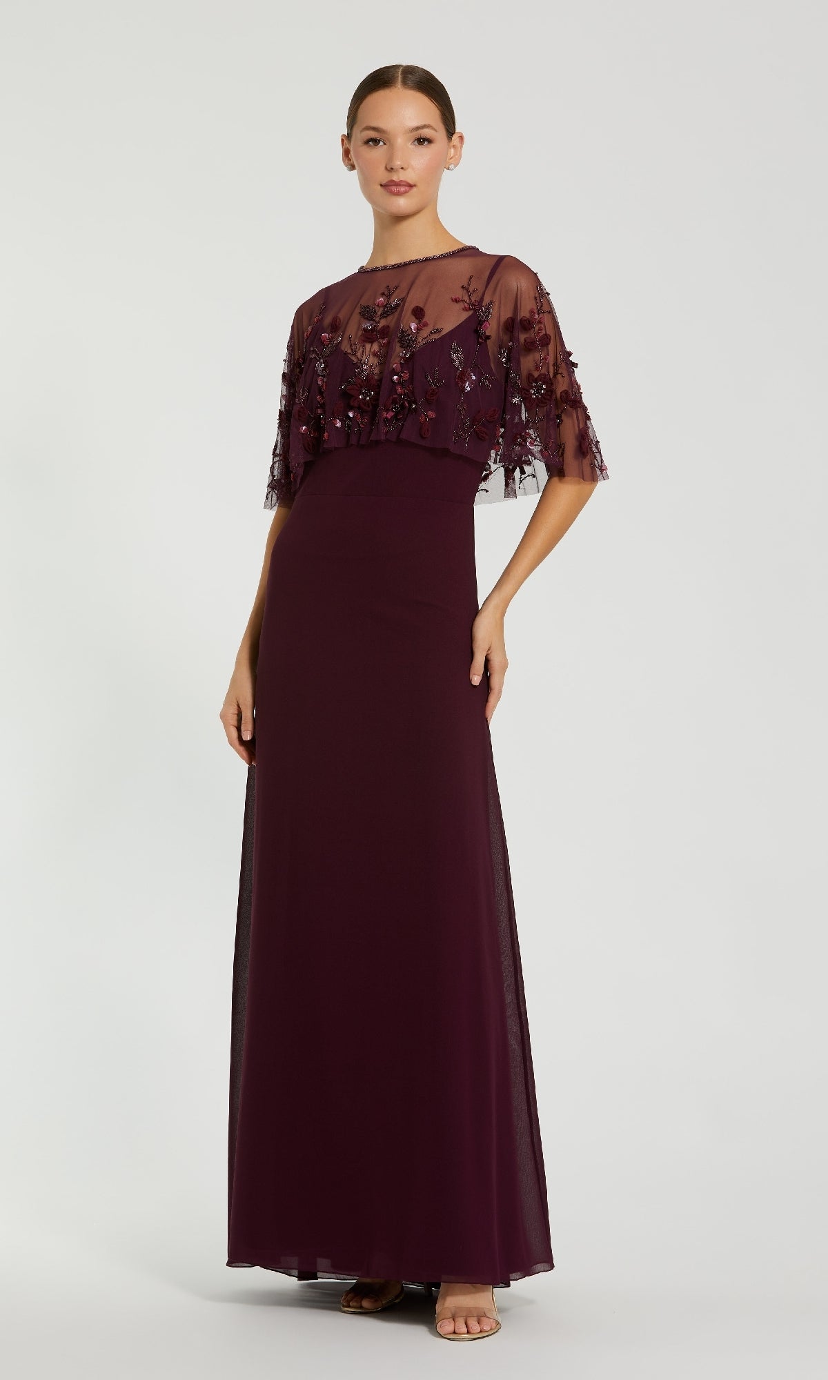 A woman wears the Mac Duggal 9229, a floor-length deep purple prom dress featuring sheer, embroidered short sleeves and a matching bodice overlay.