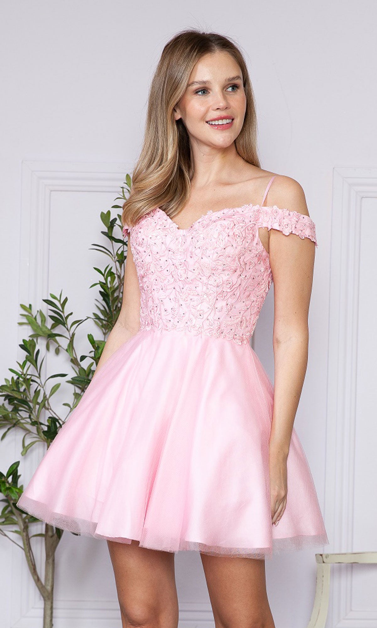 A woman stands by a white wall and green plant, wearing the Lace-Up Off-Shoulder Short Babydoll Hoco Dress9234 in light pink with lace and sequin details.