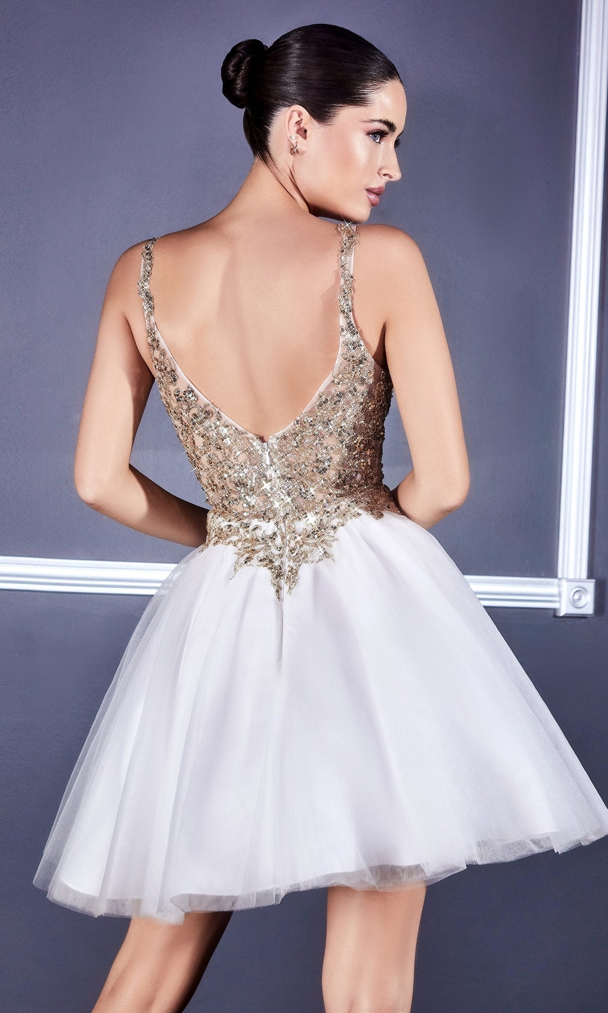 A woman stands facing away against a gray wall, wearing the Ballerina-Style Short Homecoming Dress 9239 with a gold sequin bodice, metallic-lace applique, and a short white tulle skirt.