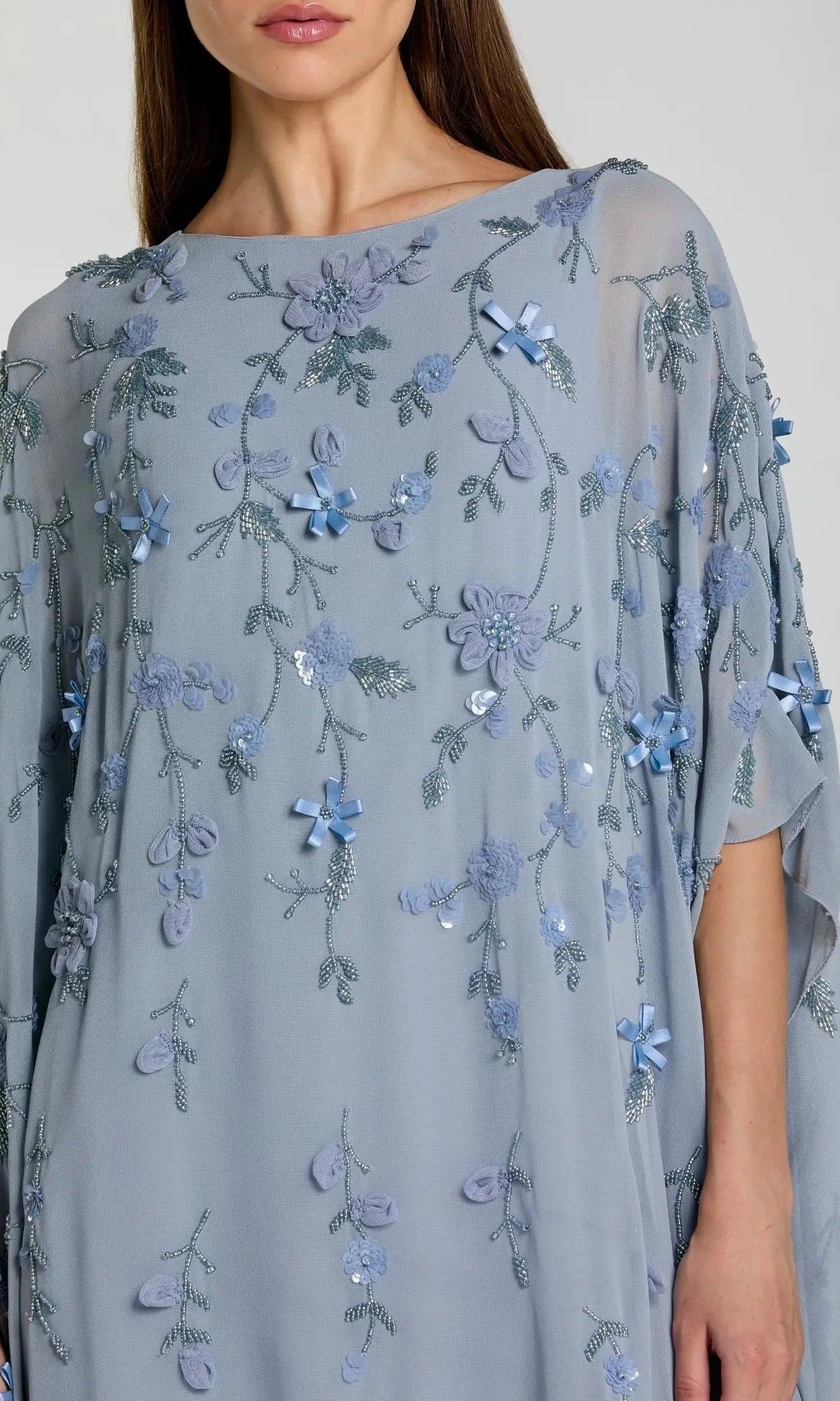 A woman wears the Trapeze Embellished Formal Gown (Mac Duggal 9242), a light blue sheer dress detailed with embroidered flowers, small blue bows, and delicate sequins for an elegant evening style.