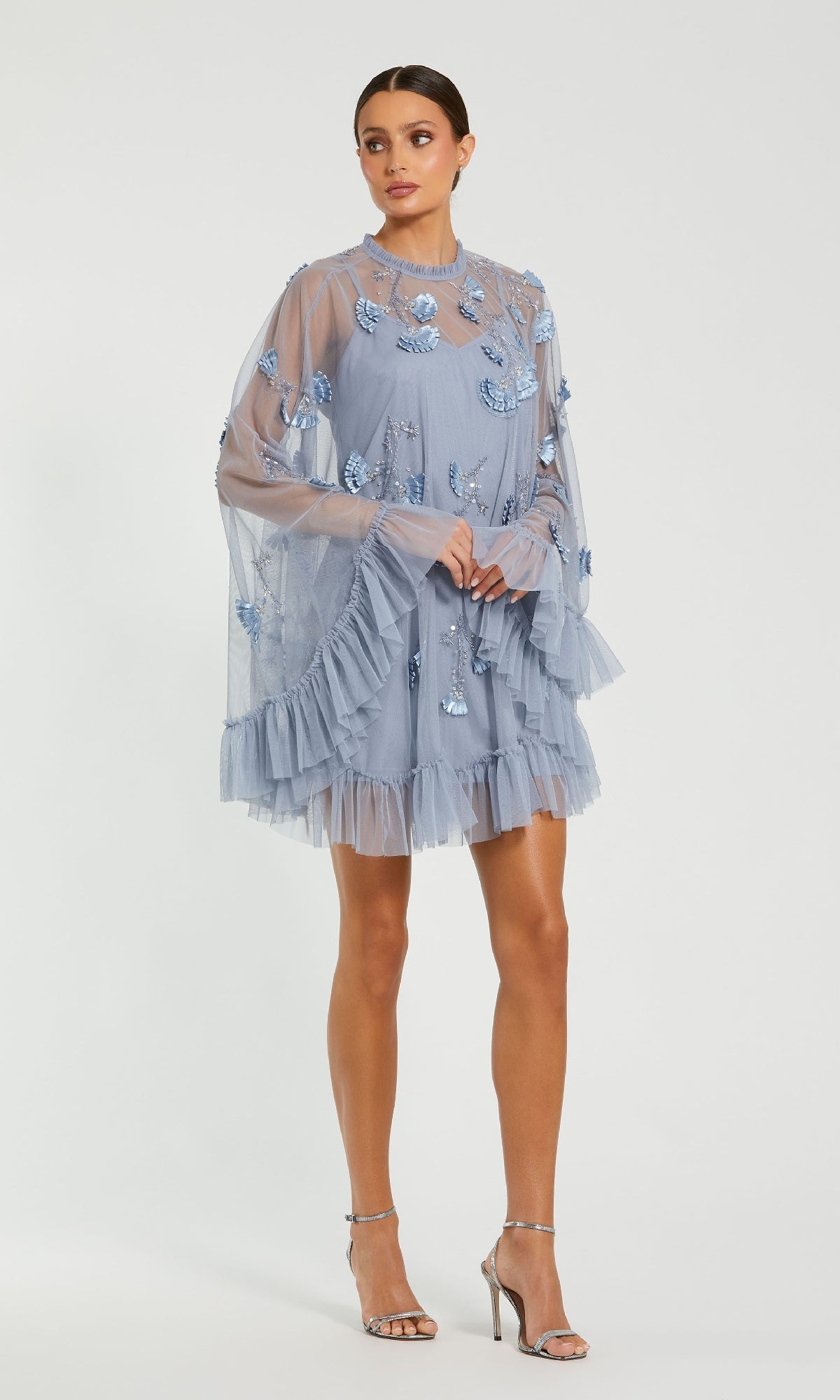 A woman wears the Mac Duggal 9257 Long Sleeve Short Hoco Dress in light blue with sheer fabric, ruffles, and floral embroidery, styled with silver high-heeled sandals.